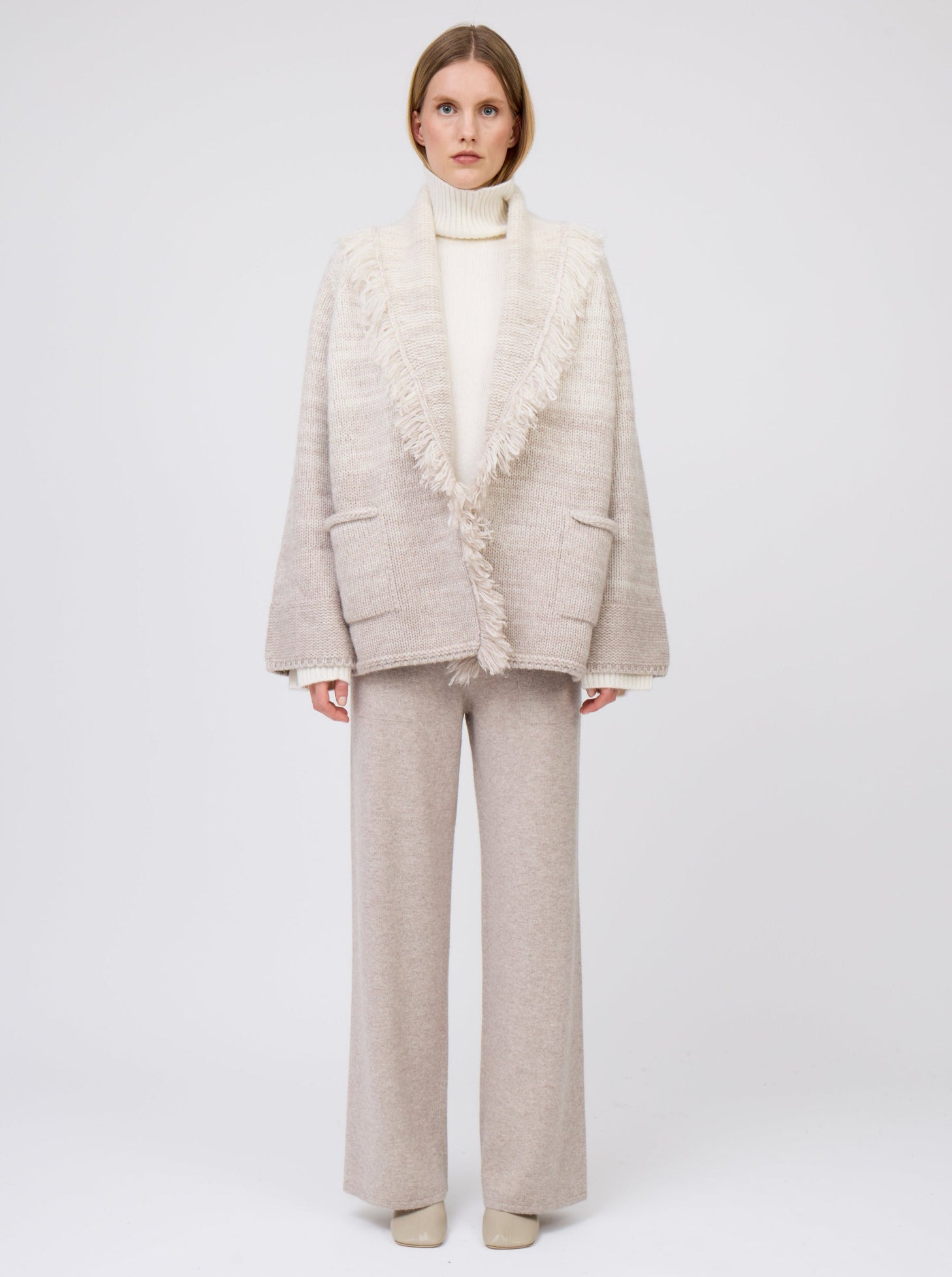 Model wears  gradient colour mix knitted cashmere jacket in white and pale beige over white cashmere roll neck sweater and beige wide leg cashmere pants.