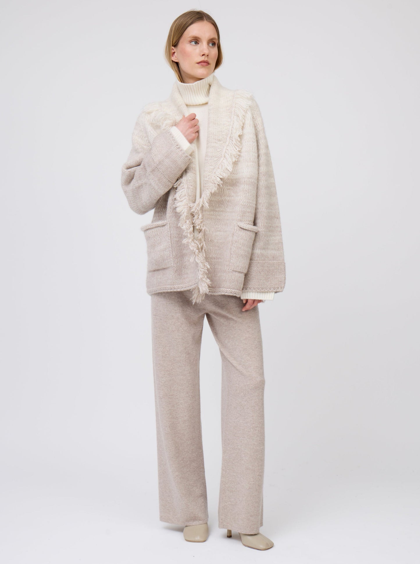 Model wears gradient colour mix knitted cashmere jacket with fringed shawl collar in white and pale beige over white cashmere roll neck sweater and beige wide leg cashmere pants.
