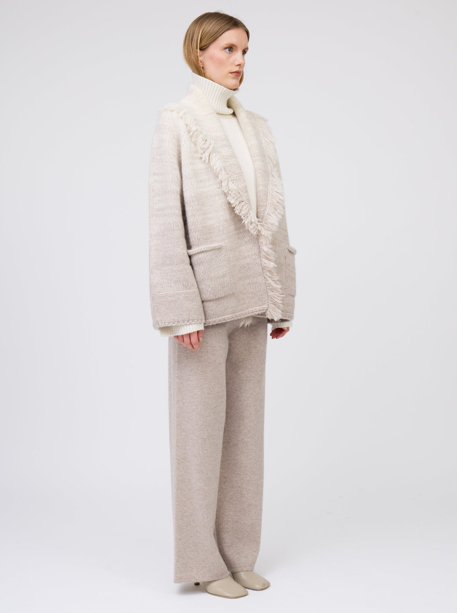 Model wears 2 colour mix knitted cashmere jacket with fringed shawl collar in white and beige over white cashmere roll neck sweater and beige wide leg cashmere pants.