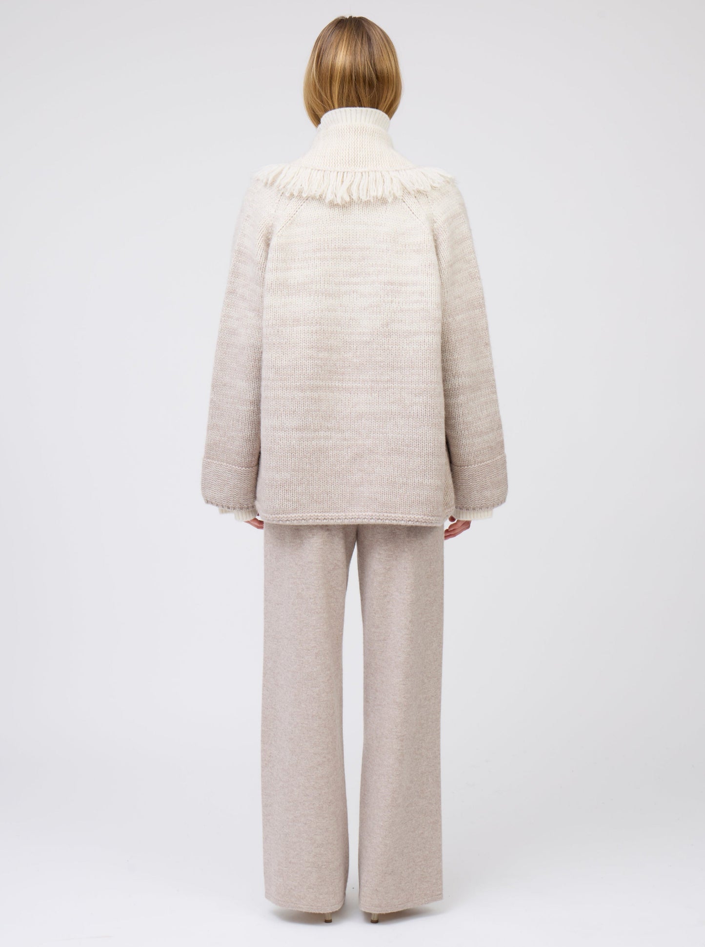Back view of model wearing gradient colour mix knitted cashmere jacket with deep shawl collar in white and pale beige over beige wide leg cashmere pants.