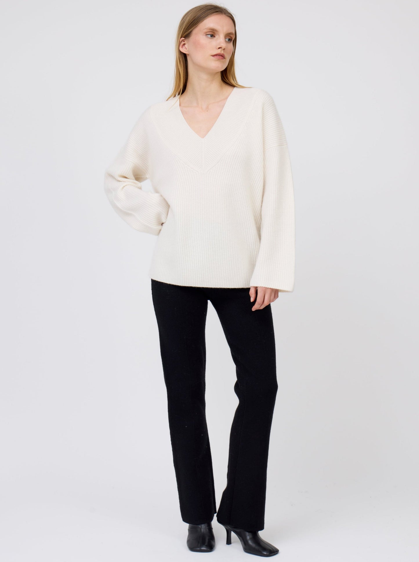 Model wears white deep V-neck ribbed knit cashmere jumper with generous long sleeves; worn with black suit pants and black boots.