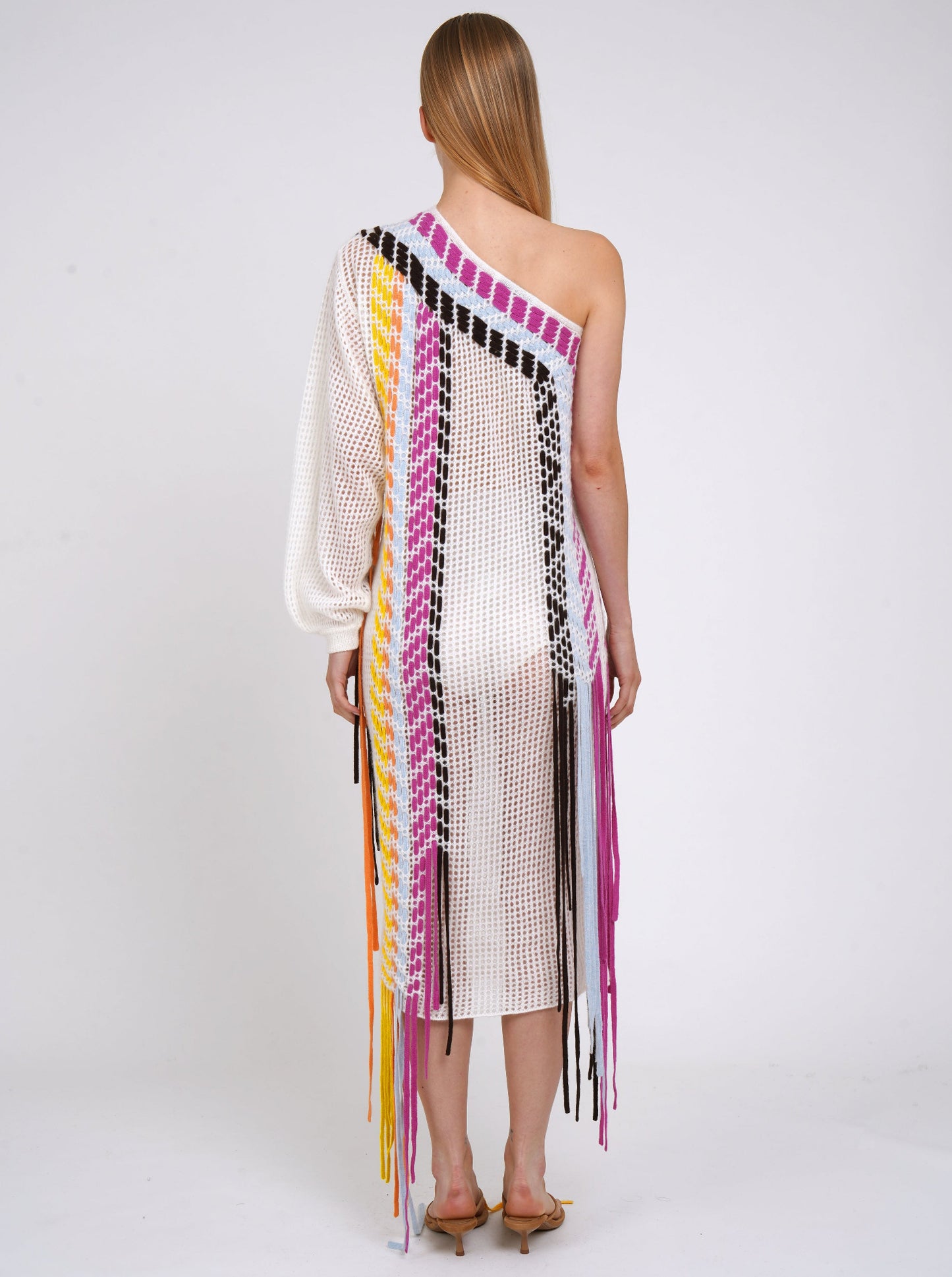 Back view white summer weight cashmere knit one shoulder shift dress with asymmetric neckline, one long sleeve, decorated with coloured woven fringes which fall to the ground.