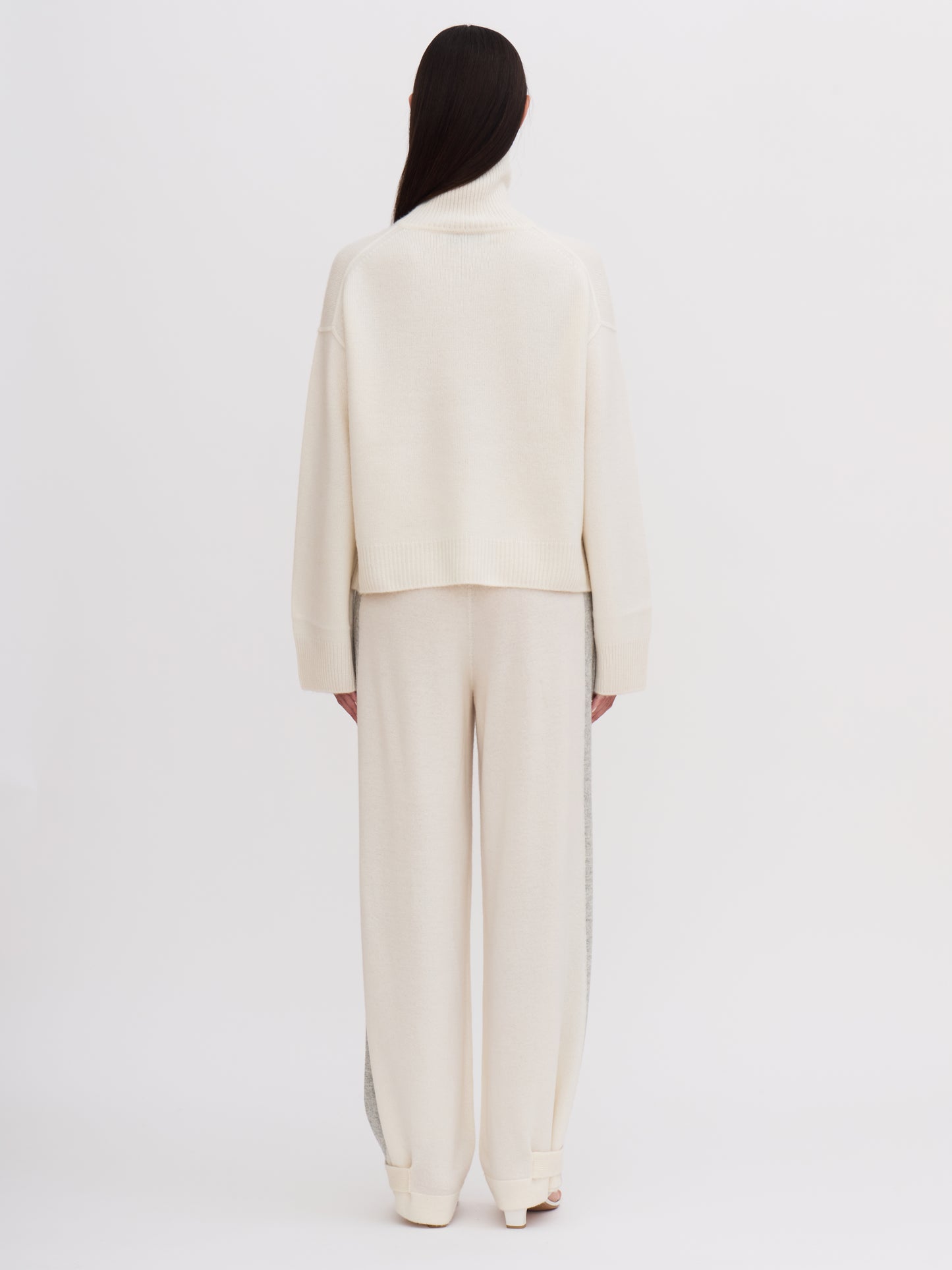 Back view of model wearing white cashmere, cropped, roll neck jumper with white wide leg pants.