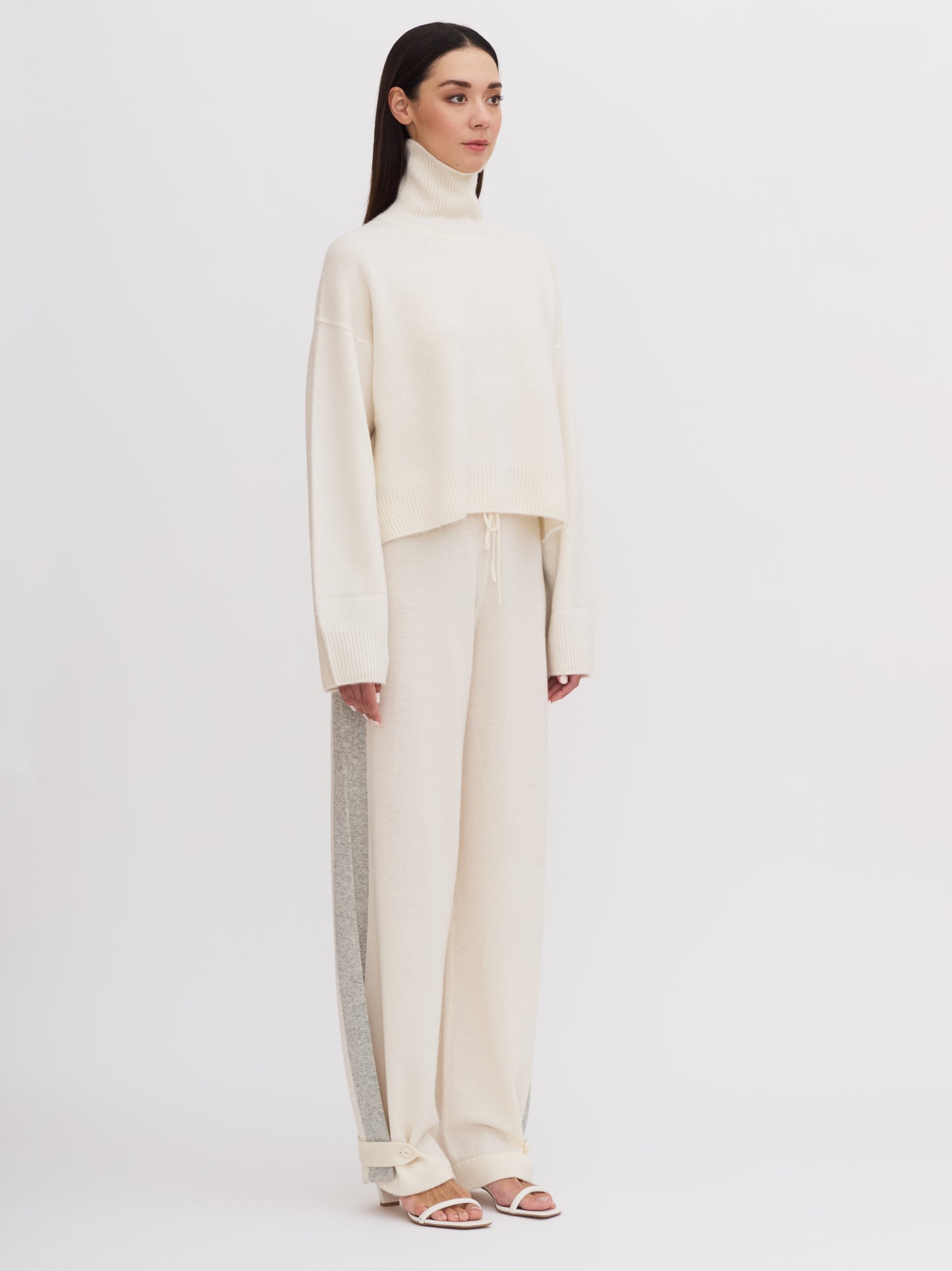 Side view of model wearing white cashmere, cropped length, roll neck jumper with wide sleeves and white wide leg pants.