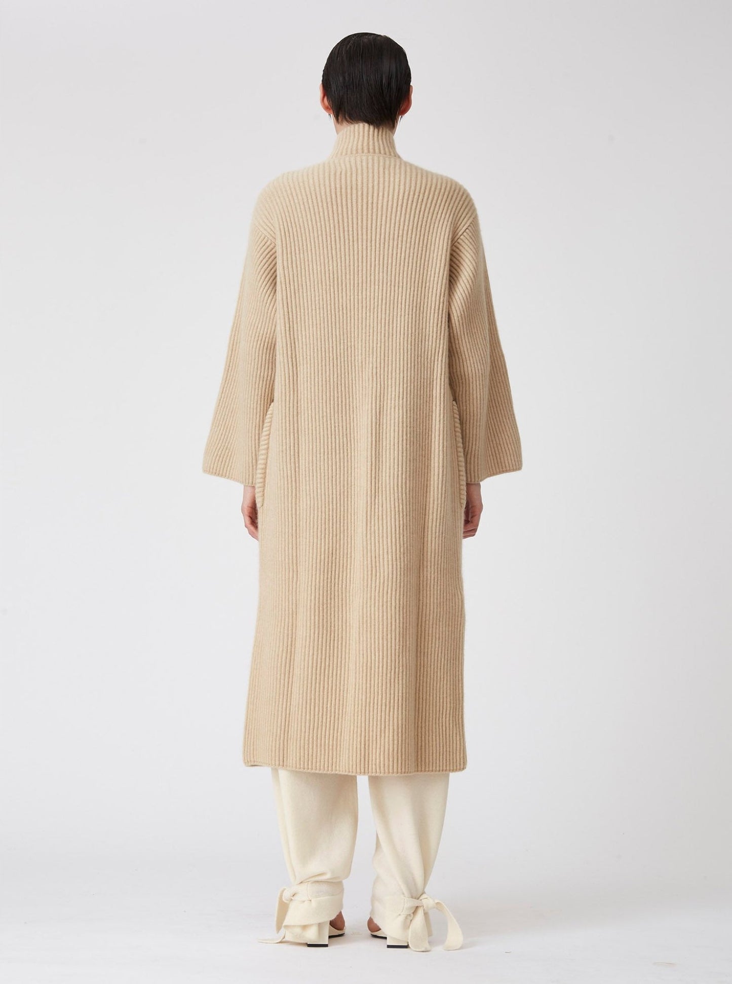 Back view of model wearing a beige midi cardigan coat in ribbed cashmere knit. Straight cut with a relaxed fit, deep patch pockets and generous sleeves which can be rolled back.