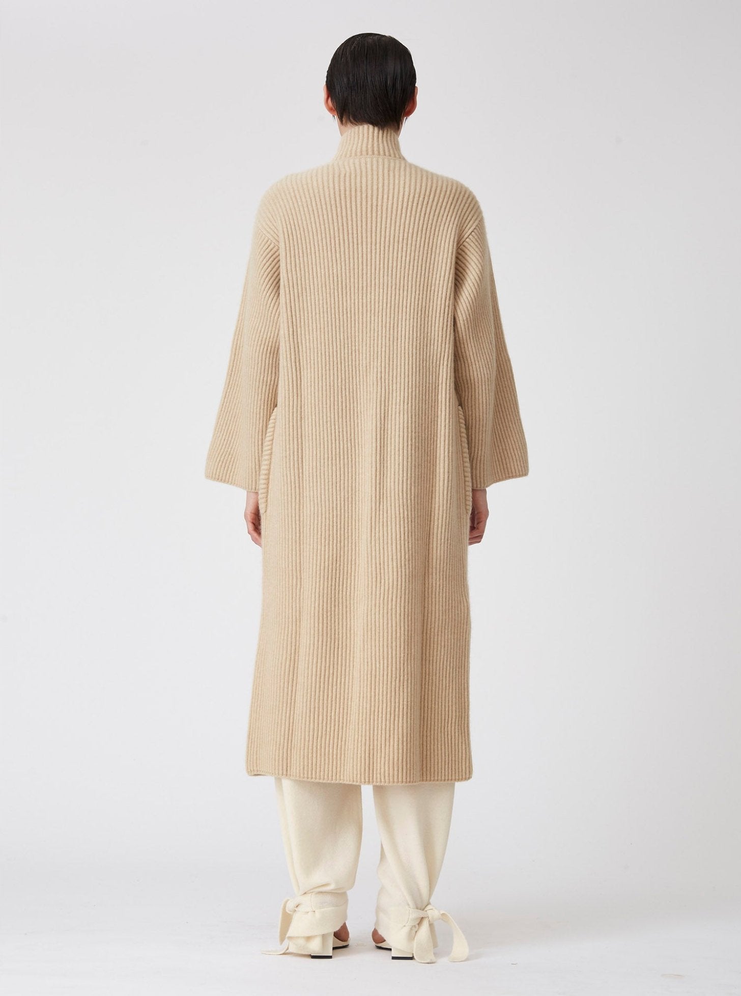 Back view of model wearing a beige midi cardigan coat in ribbed cashmere knit. Straight cut with a relaxed fit, deep patch pockets and generous sleeves which can be rolled back.