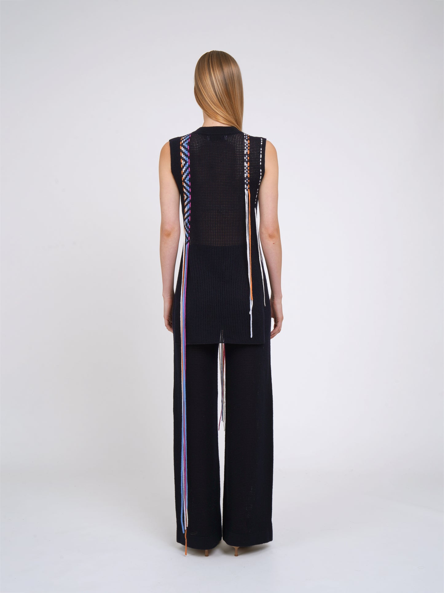 back view dark navy net knit pants worn with light weight cashmere knit sleeveless tunic decorated with woven coloured fringes.