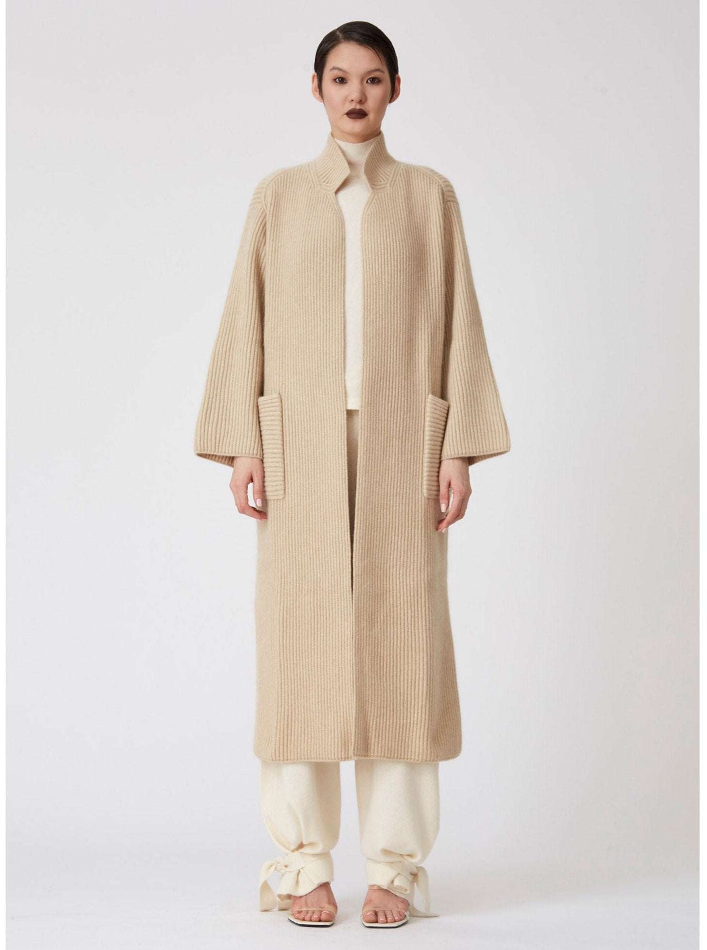 Front view of model wearing a beige Midi cardigan coat in ribbed cashmere knit. Straight cut with a relaxed fit, deep patch pockets and generous sleeves which can be rolled back. 