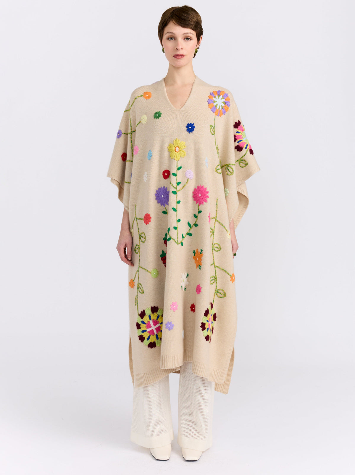 Front straight view of model wearing midi length beige cashmere kaftan with V neck, open sides and embroidered coloured flowers worn over white cashmere knit pants.