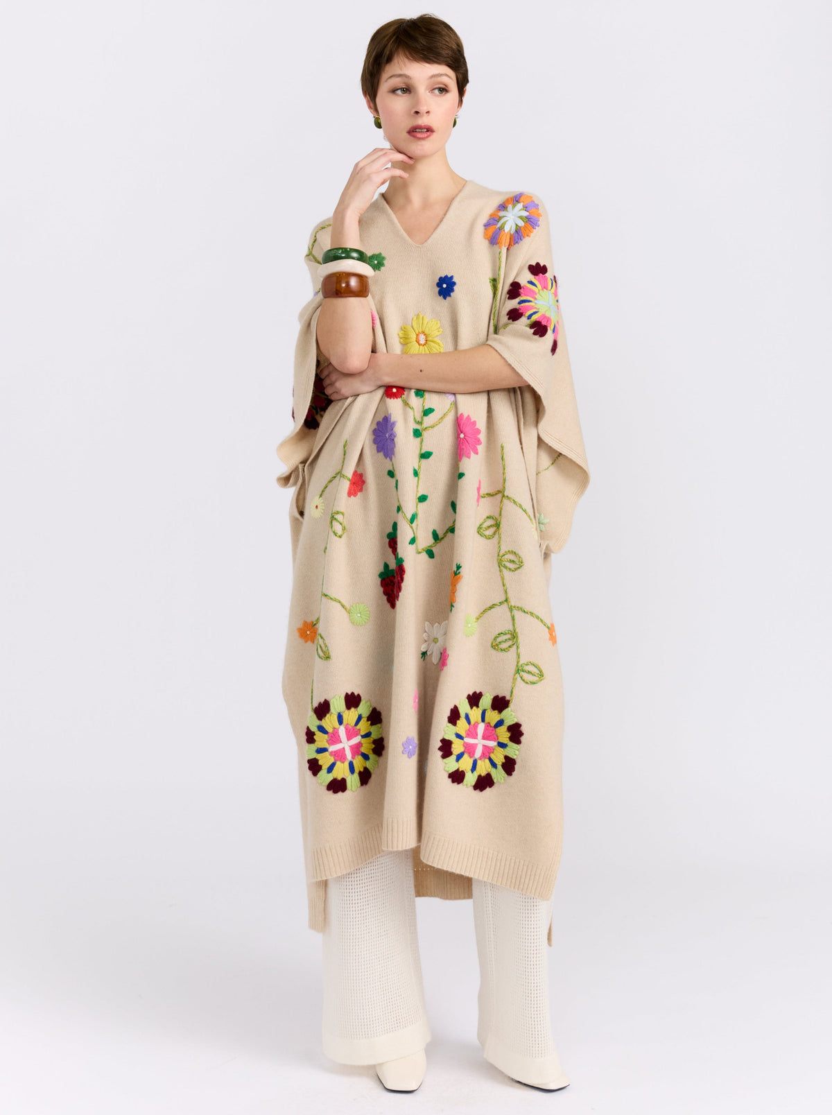 Posed model wearing midi length beige cashmere kaftan with V neck, open sides and embroidered coloured flowers worn over white cashmere knit pants and styled with large colour bangles.