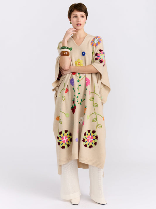 Posed model wearing midi length beige cashmere kaftan with V neck, open sides and embroidered coloured flowers worn over white cashmere knit pants and styled with large colour bangles.