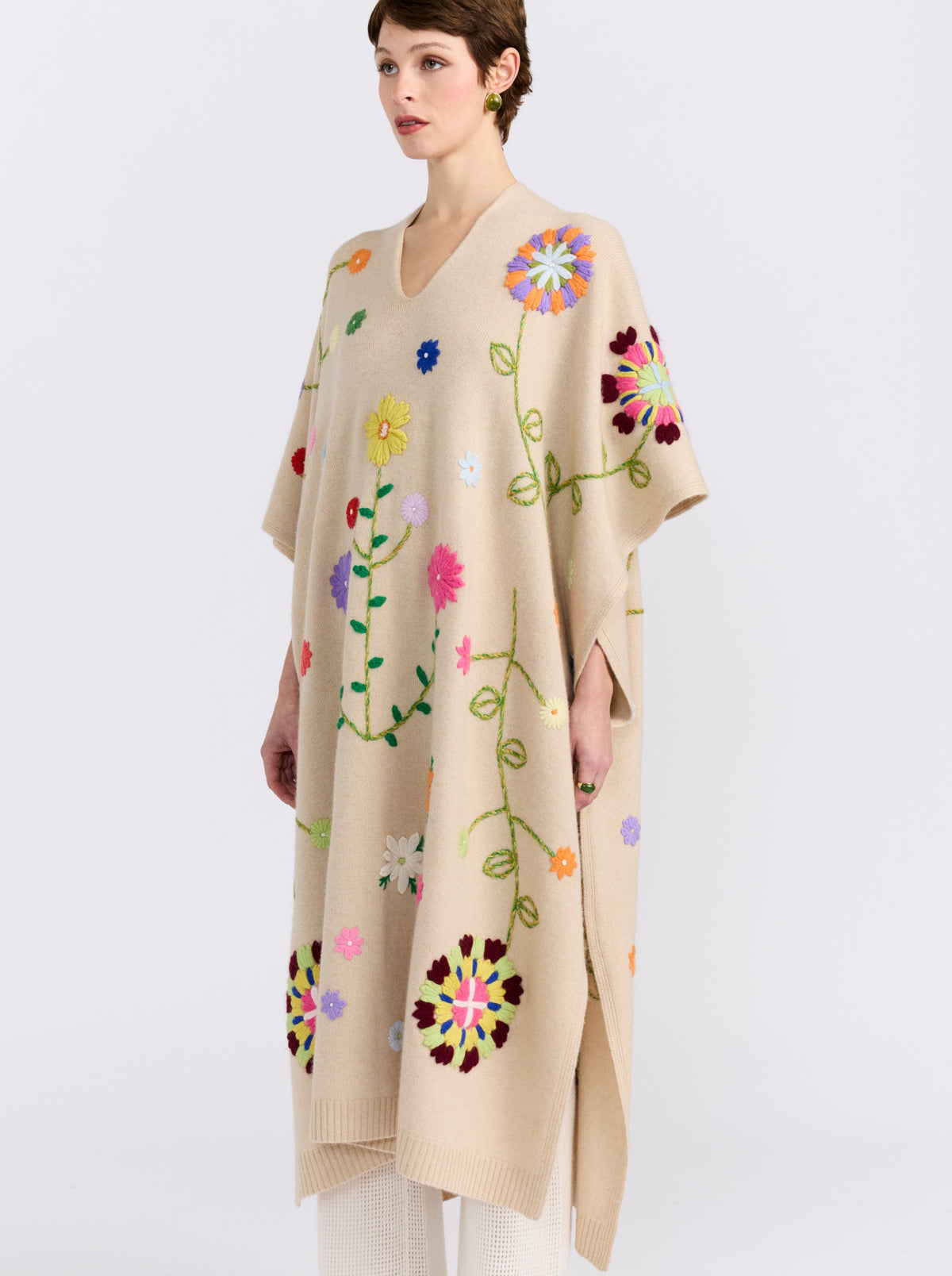 Side view of model wearing midi length beige cashmere kaftan with V neck, open sides and embroidered coloured flowers worn over white cashmere knit pants.