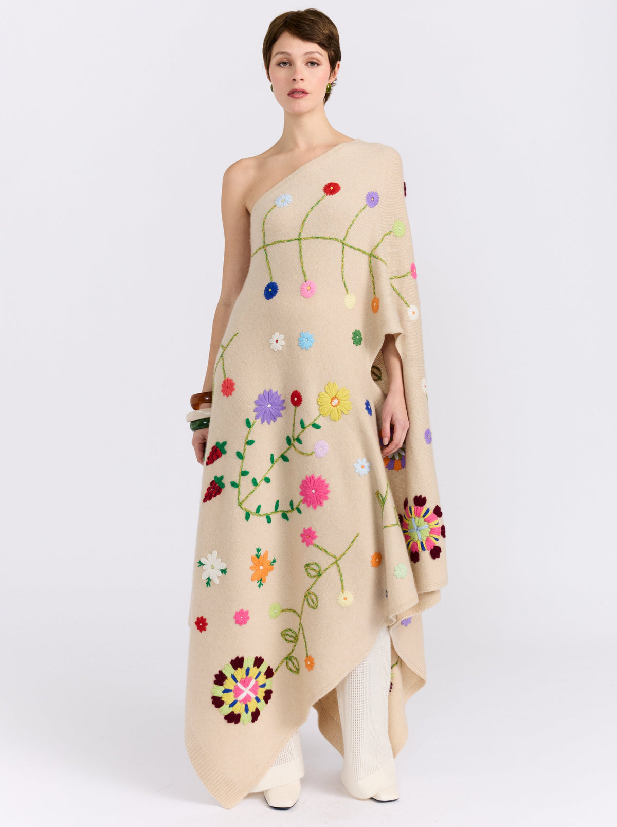 Front view of model wearing full length beige cashmere asymmetric kaftan with embroidered coloured flowers worn as a one shoulder dress over white knit pants.