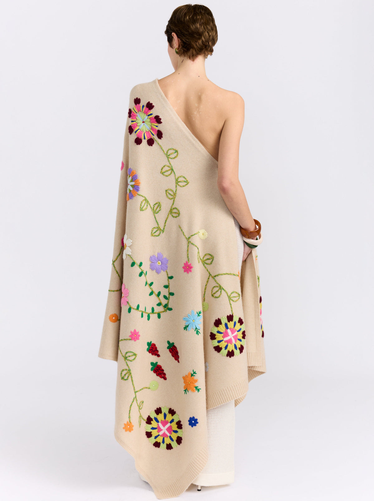 model's back view wearing full length beige cashmere kaftan with embroidered coloured flowers worn as a one shoulder dress.