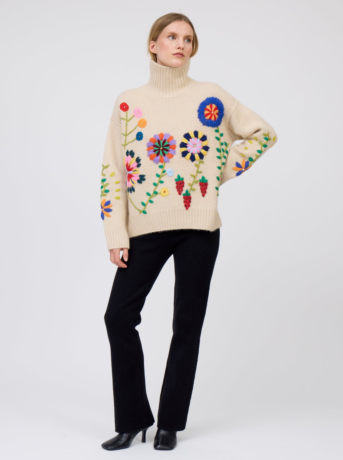 Model wearing beige oversized roll neck sweater adorned with brightly coloured hand embroidered flowers; worn with black cashmere suit pants.