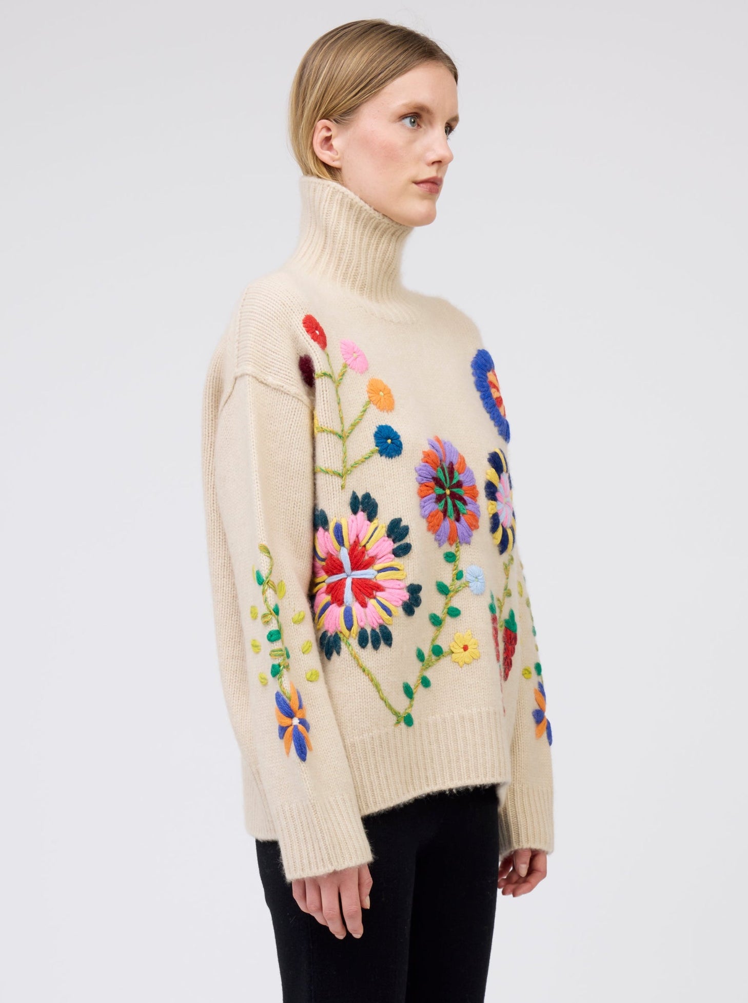 Close up of model wearing beige oversized roll neck sweater adorned with bold design of brightly coloured hand embroidered flowers.