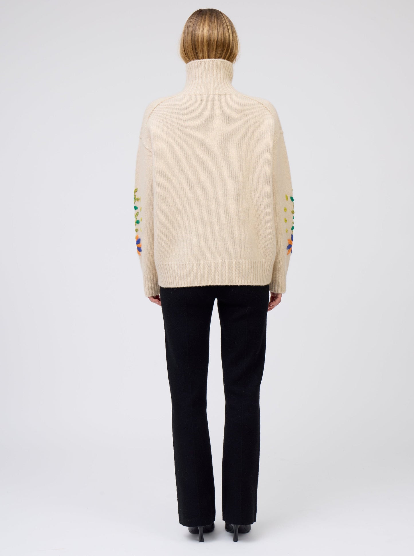Back view of model wearing beige cashmere oversized roll neck sweater with some embroidery visible on sleeves; worn with black cashmere suit pants.
