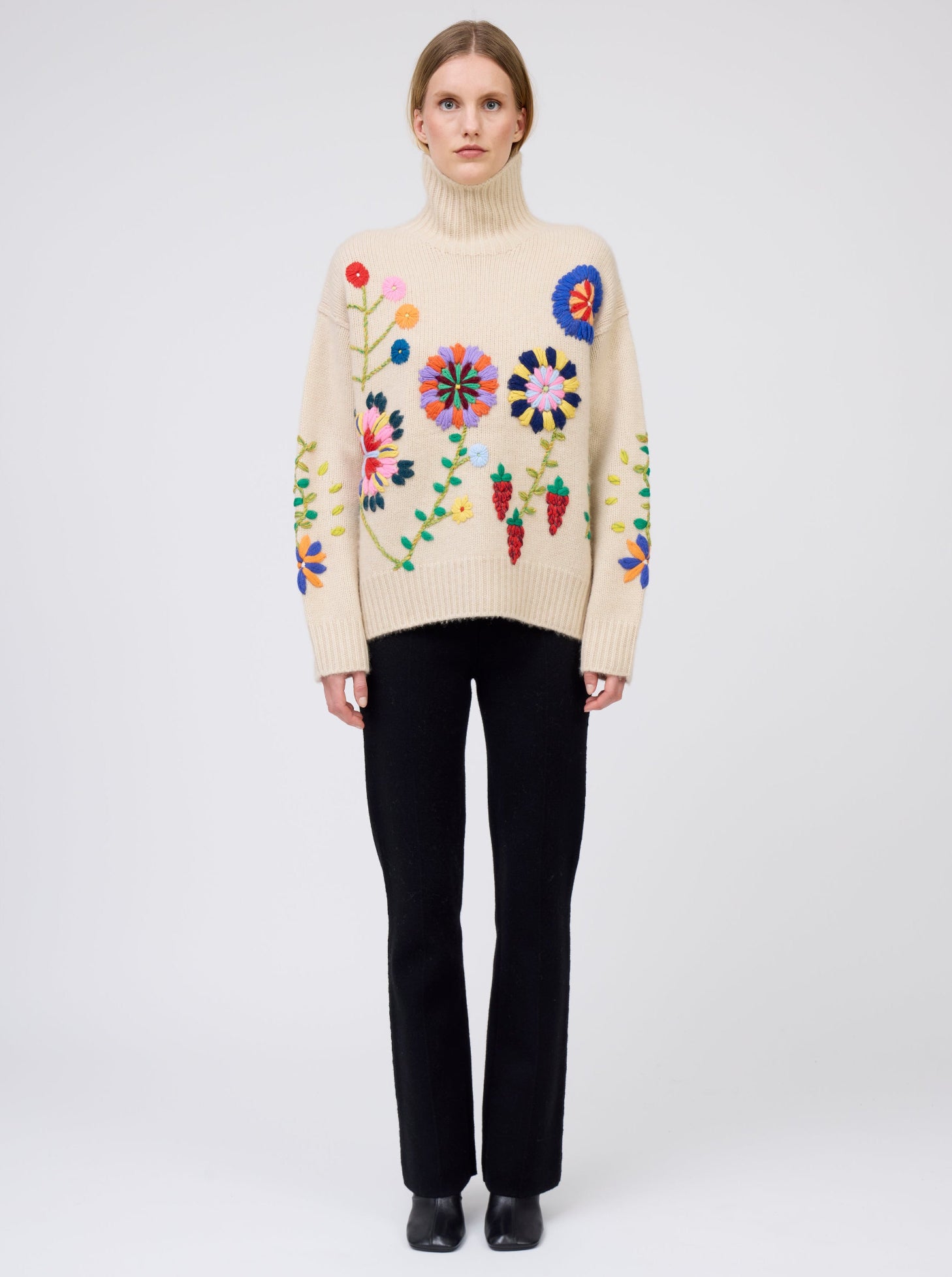 Front view of model wearing beige cashmere oversized roll neck sweater adorned with brightly coloured hand embroidered flowers; worn with black cashmere suit pants.