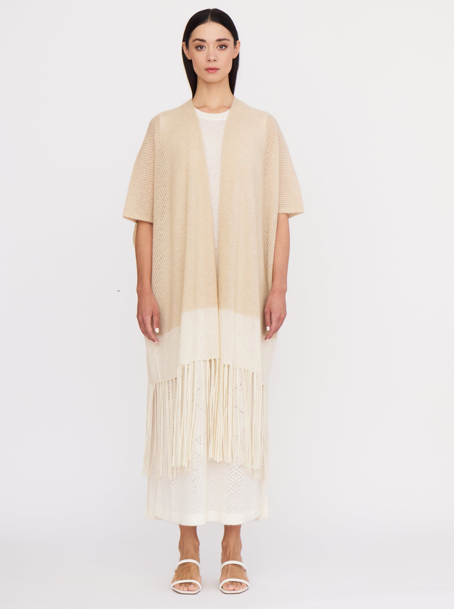 Front view of model wearing light weight cashmere knit cover up in beige and white with elbow length sleeves and fringed hem, over white cashmere dress.