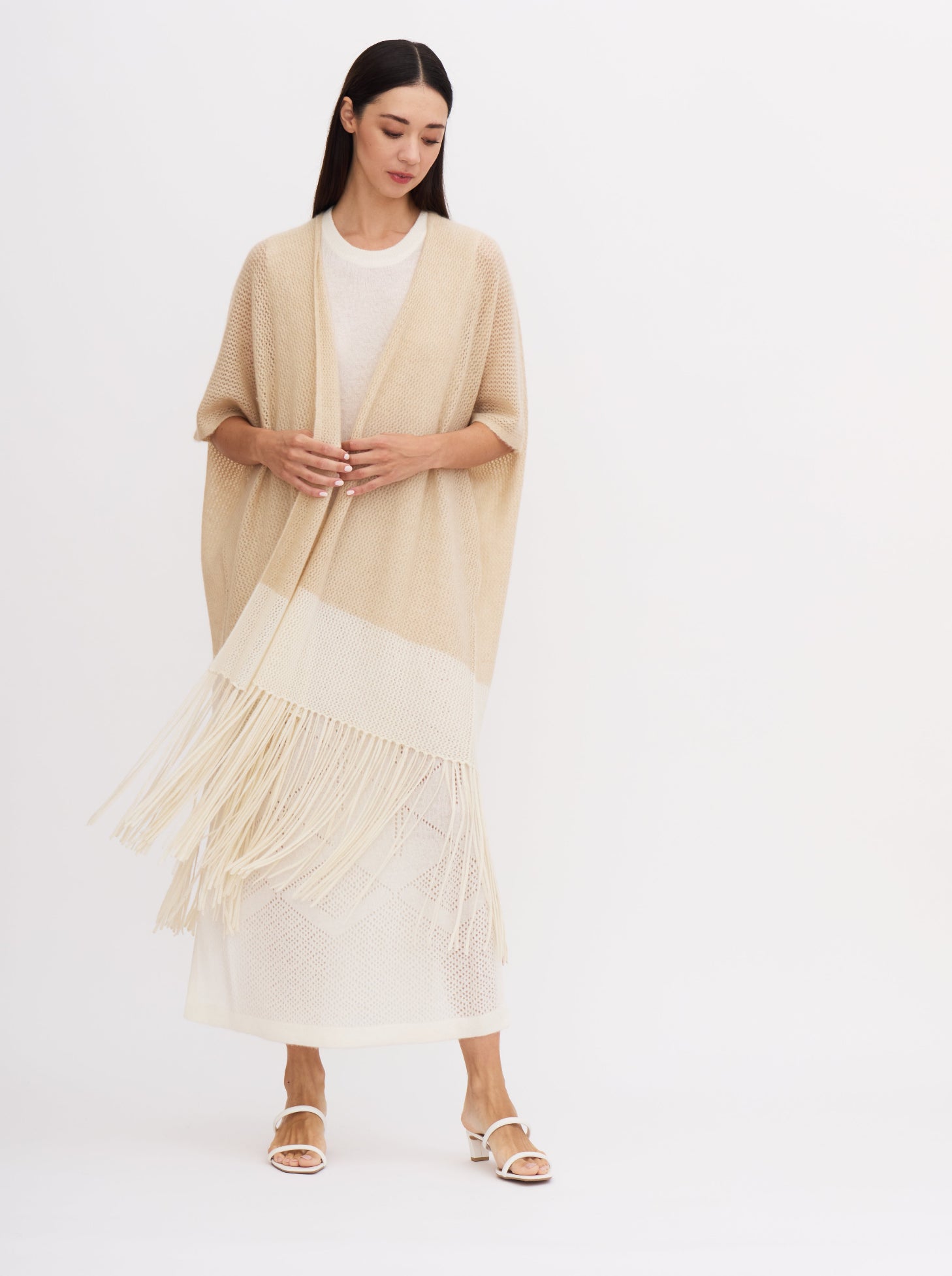 Model wears relaxed fit, summer cashmere knit cover up in beige and white with elbow length sleeves and fringe, over a white cashmere dress, showing the sway movement of the fringed hem.