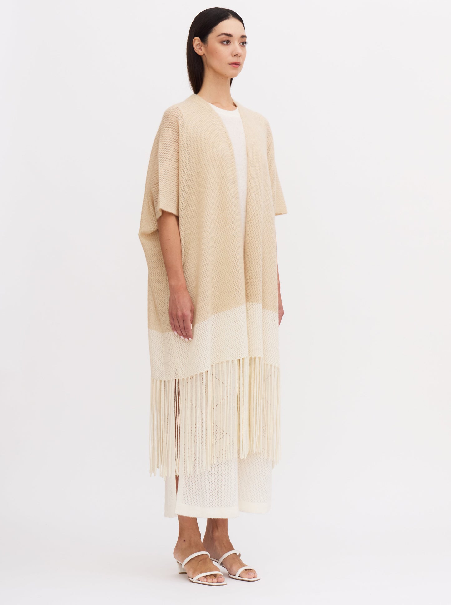 Model wears loose-fitting, summer-weight cashmere net knit cover up in beige and white with elbow length sleeves and fringed hem, over white cashmere dress.