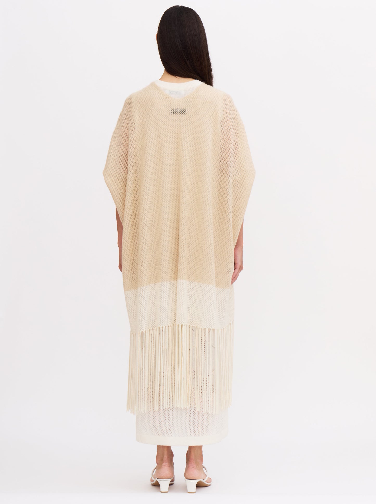 Back view of model wearing loose-fitting, light-weight cashmere knit cover up in beige and white with elbow length sleeves and fringed hem, over white cashmere dress.