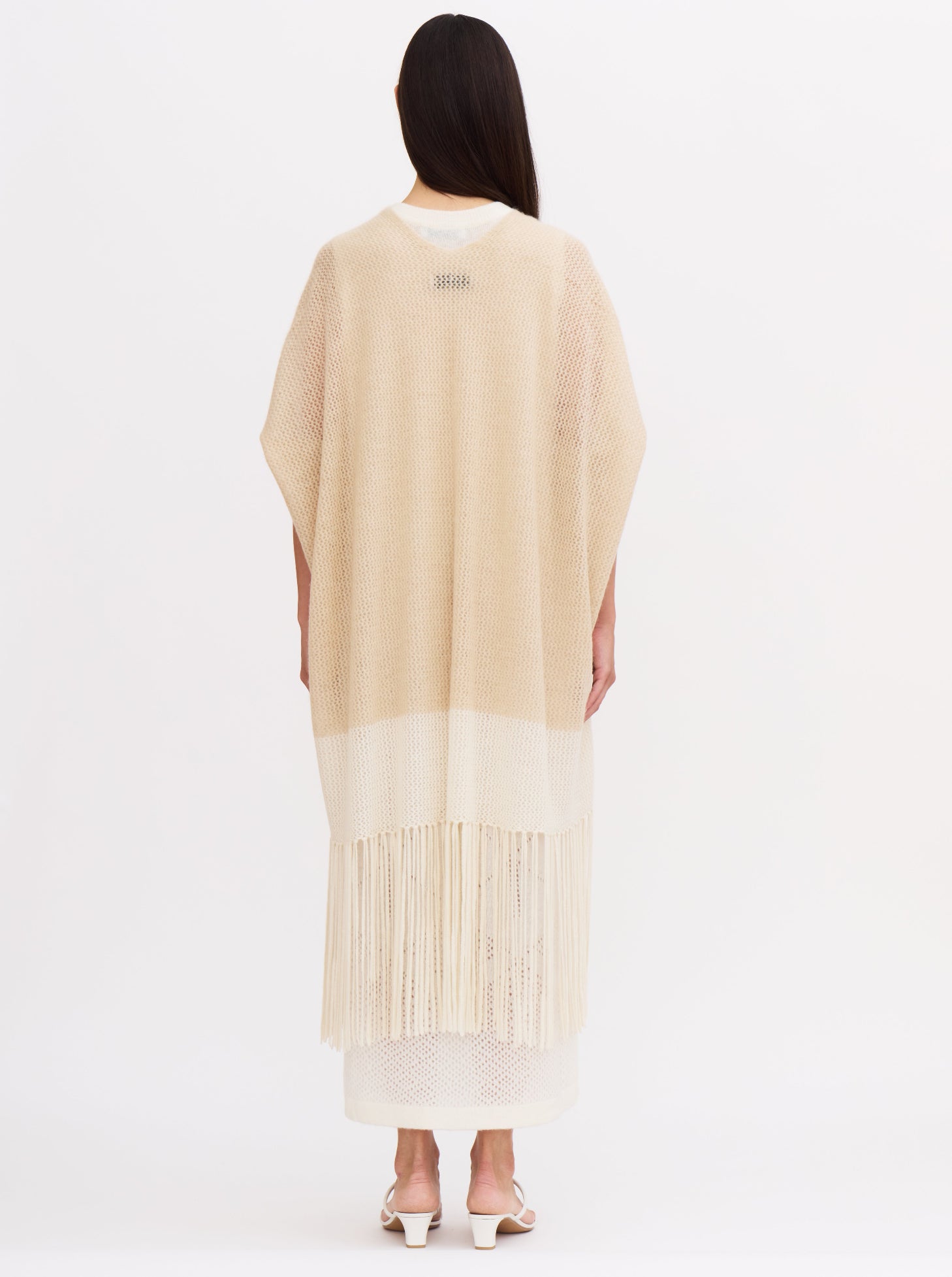 Back view of model wearing loose-fitting, light-weight cashmere knit cover up in beige and white with elbow length sleeves and fringed hem, over white cashmere dress.