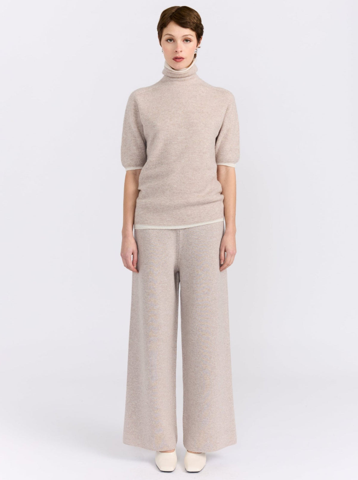 Model stands against white studio background wearing beige knit turtleneck sweater with short sleeves and matching wide-leg trousers.
