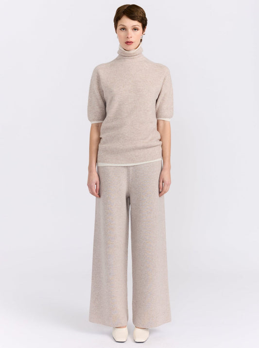 Model stands against white studio background wearing beige knit turtleneck sweater with short sleeves and matching wide-leg trousers.