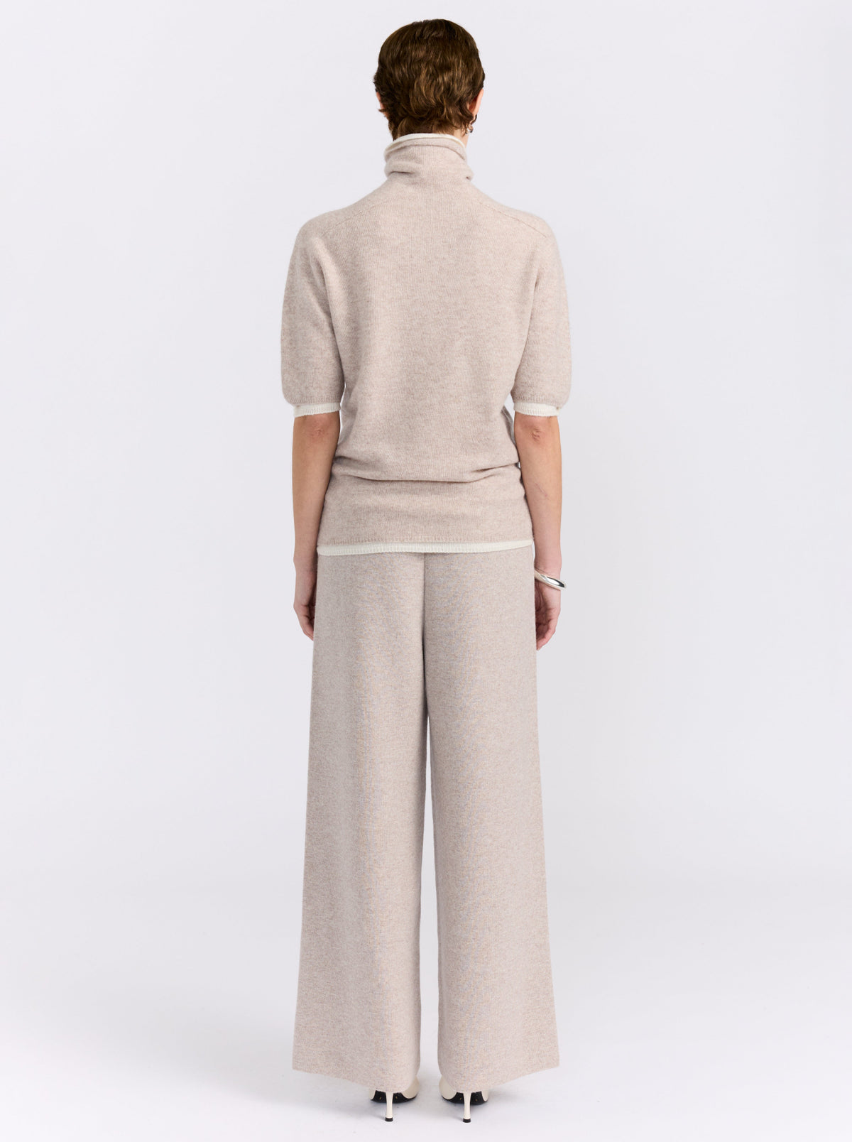 Back view model wearing beige knit turtleneck sweater with short sleeves and matching wide-leg trousers in a white background studio.