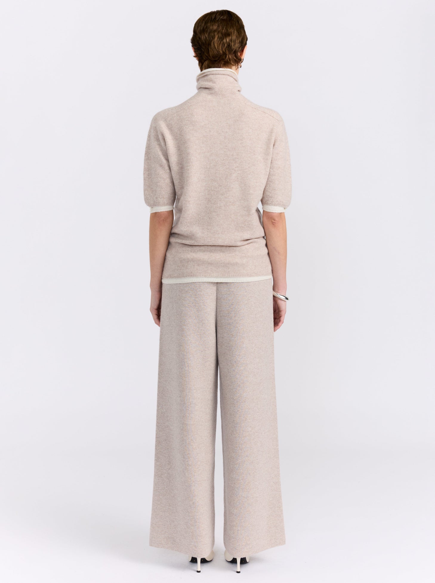 Back view model wearing beige knit turtleneck sweater with short sleeves and matching wide-leg trousers in a white background studio.