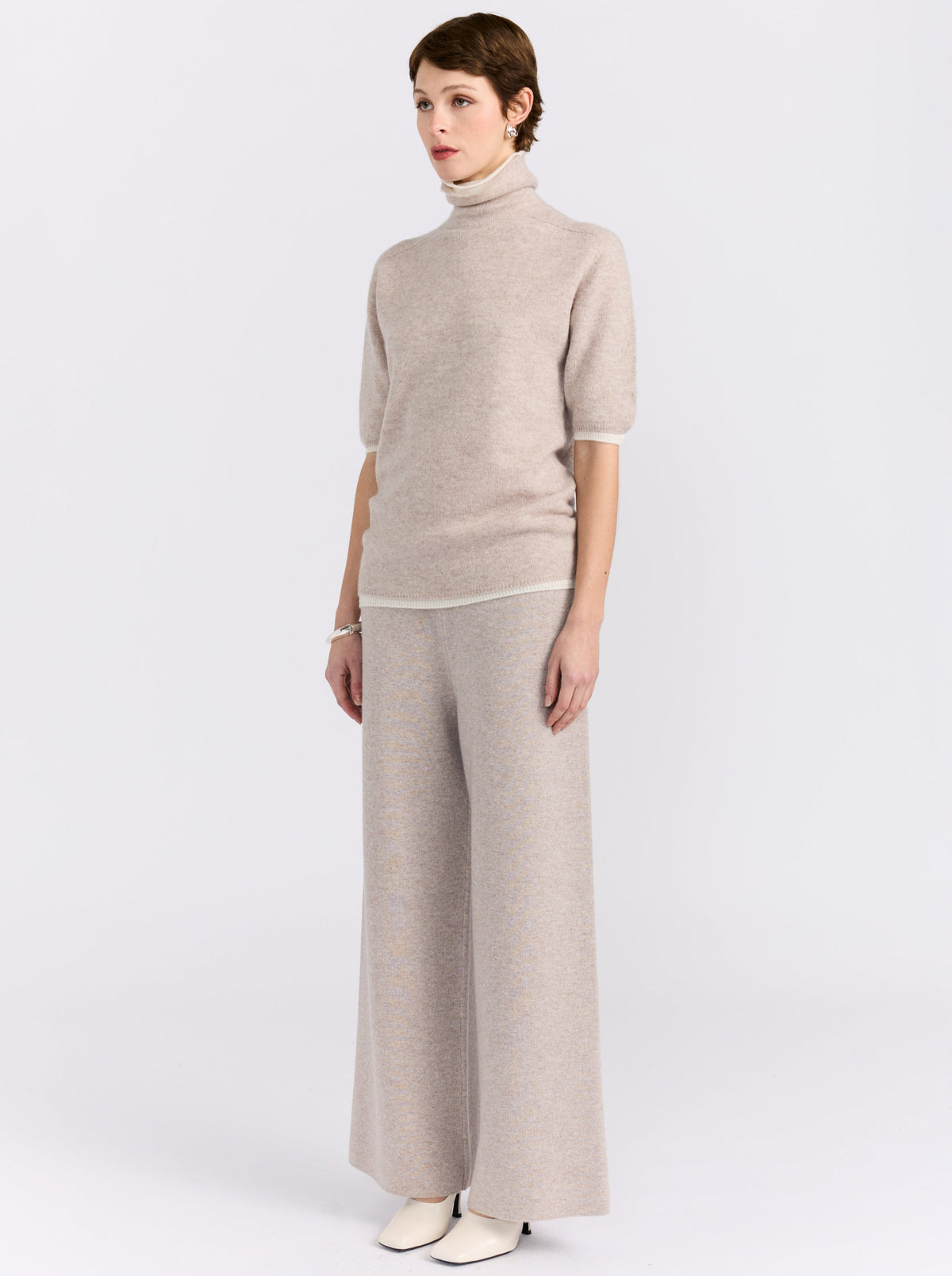 Full length side view of model wearing beige knit turtleneck short sleeve sweater and matching wide-leg trousers in a white background studio.