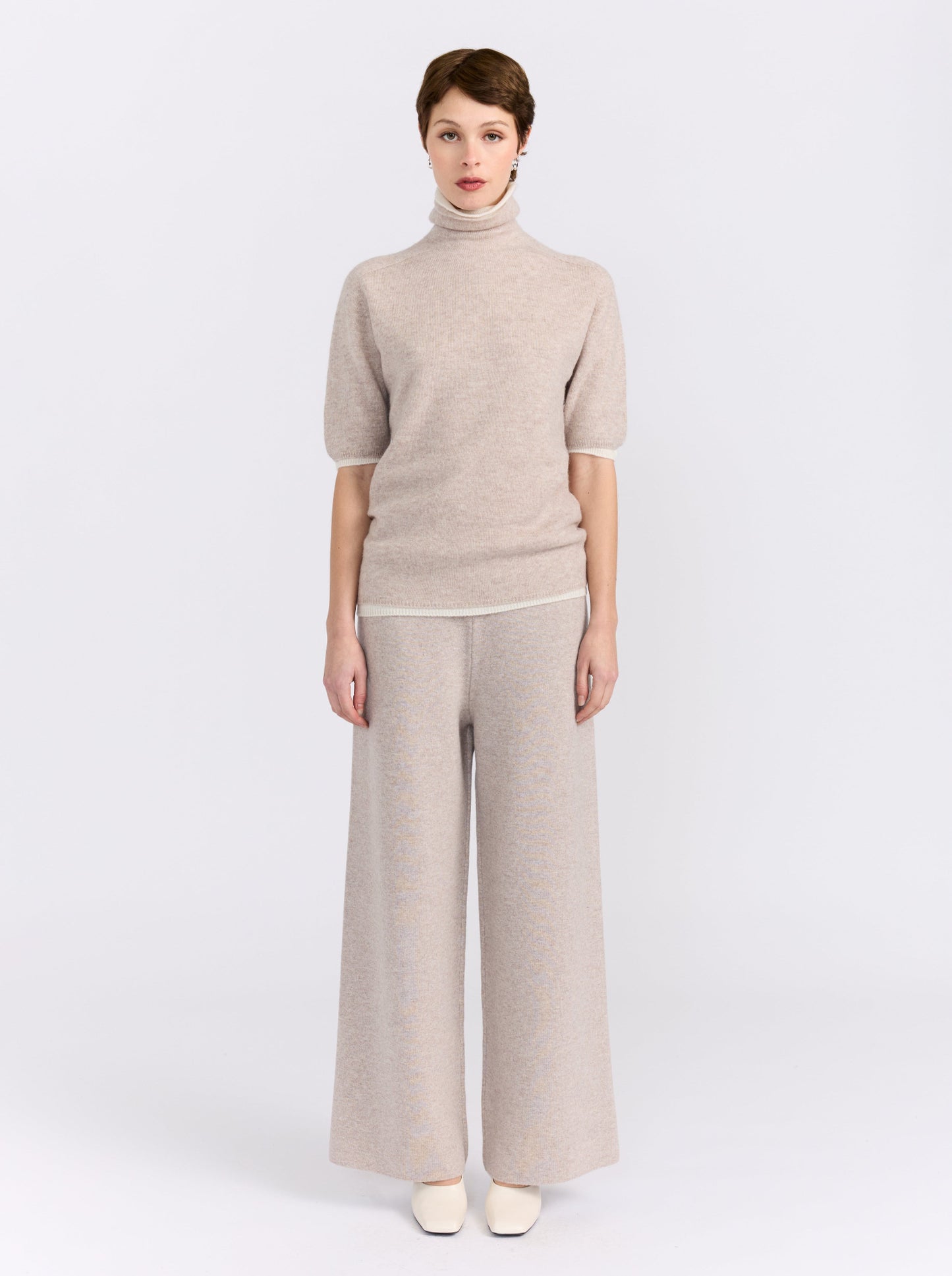 A model stands against a light gray studio background wearing a light beige knit turtleneck sweater with short sleeves and matching wide-leg trousers.