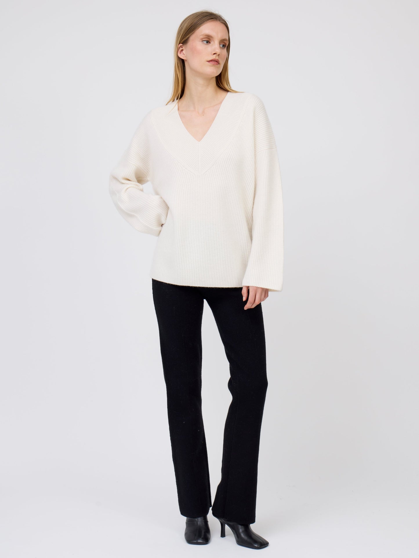 Full length front view model wearing black long tailored pants, with with slight boot cut flare, with cream V neck loose sweater and and high heel black ankle boots