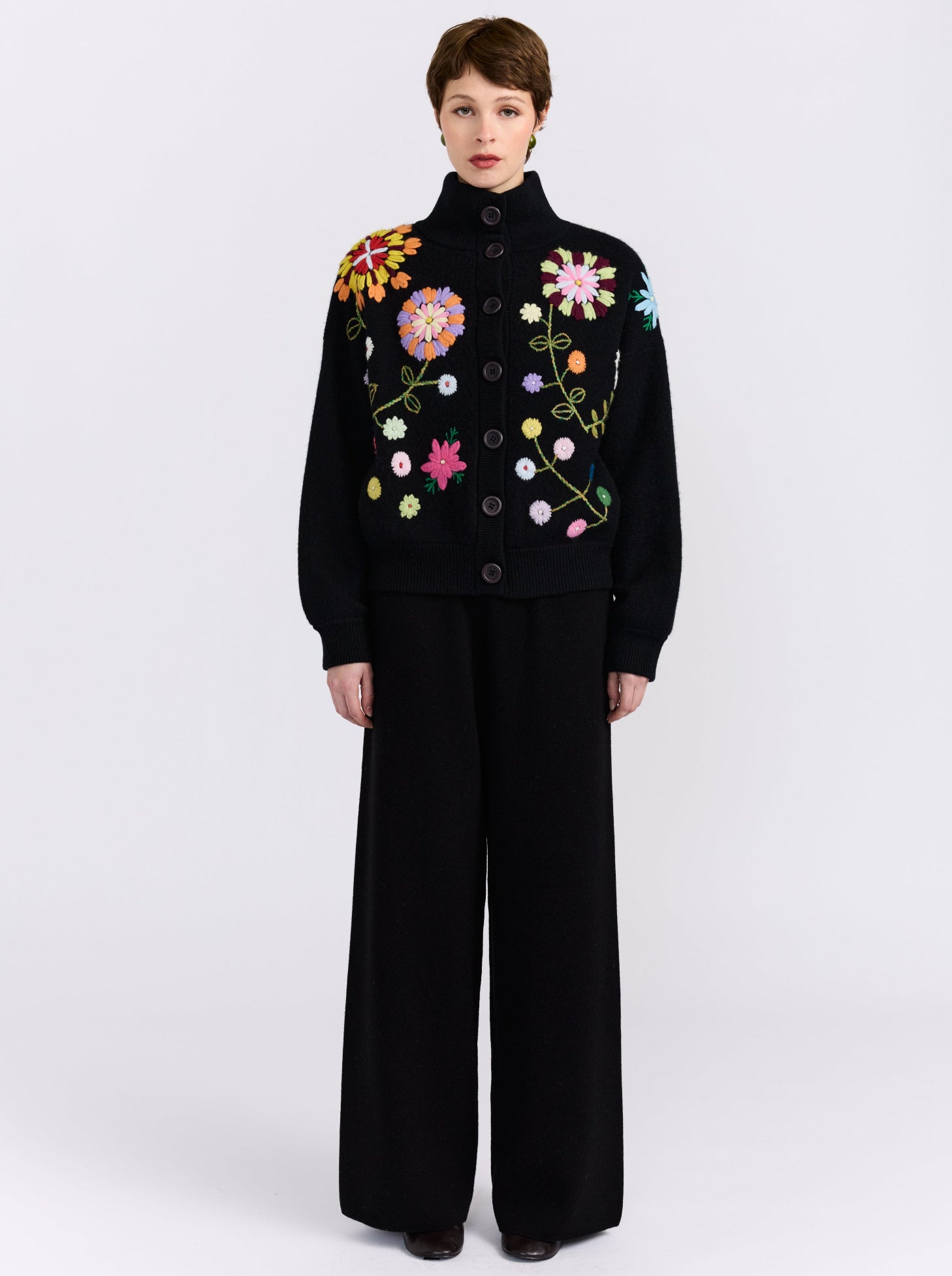 A model stands against a light gray studio background wearing a black funnel-neck bomber jacket with embroidered floral details, paired with wide-leg black trousers.