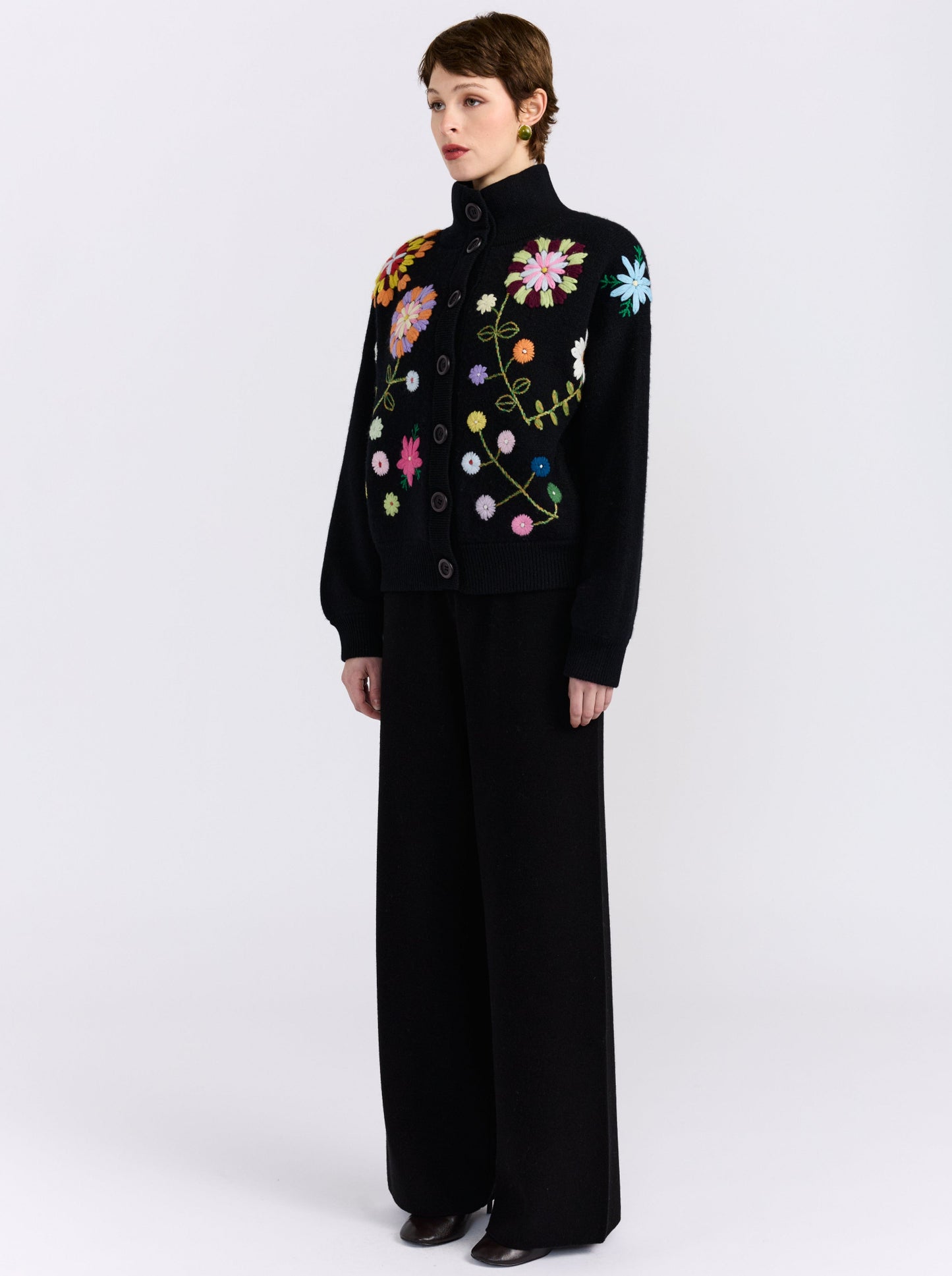 A model wearing a black funnel-neck black knit bomber jacket with embroidered floral details, paired with wide-leg black trousers, against a light gray studio background.