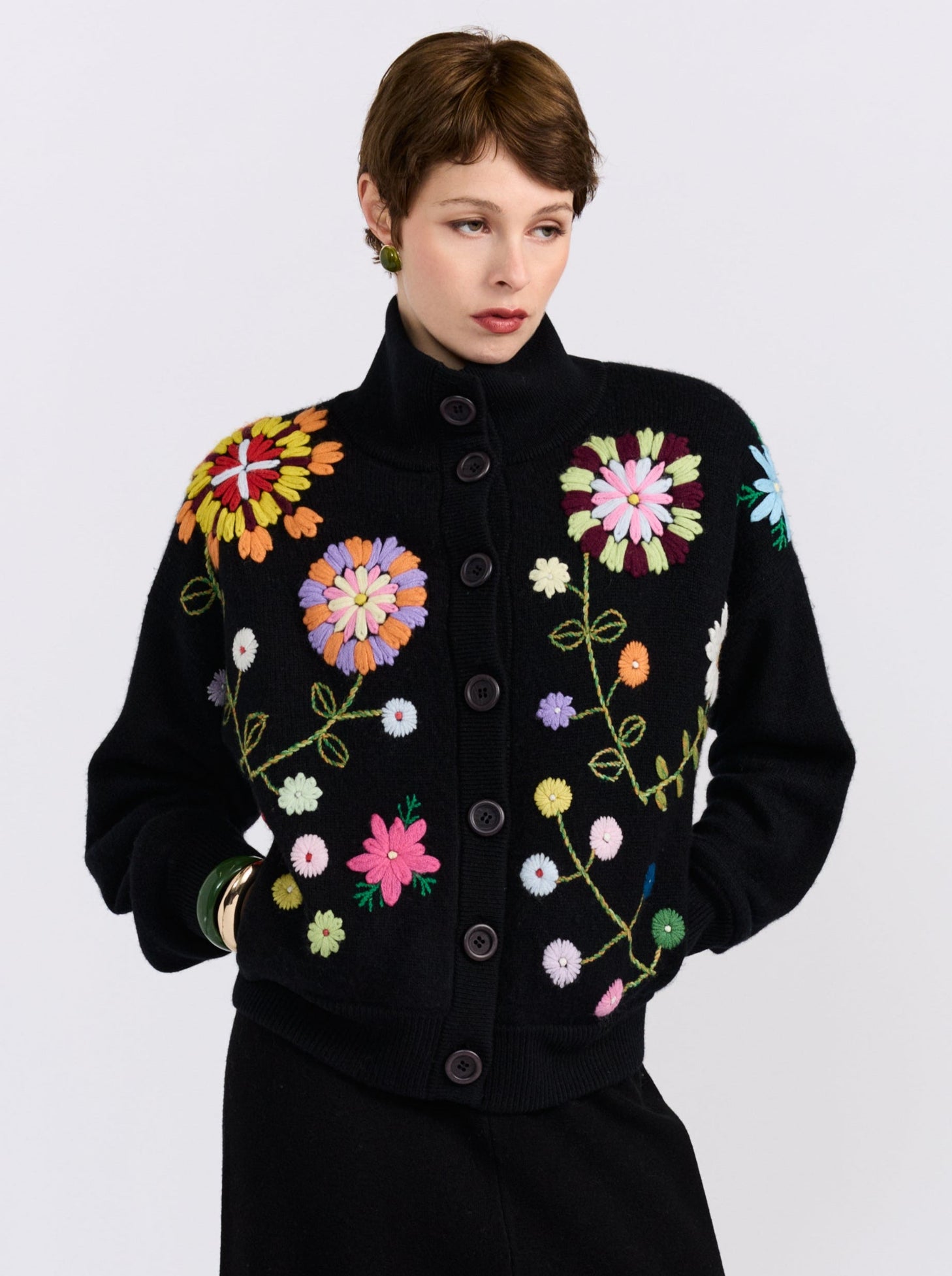 A model wearing a black knit cardigan sweater with colorful embroidered flowers and green stems, set against a plain light gray background.