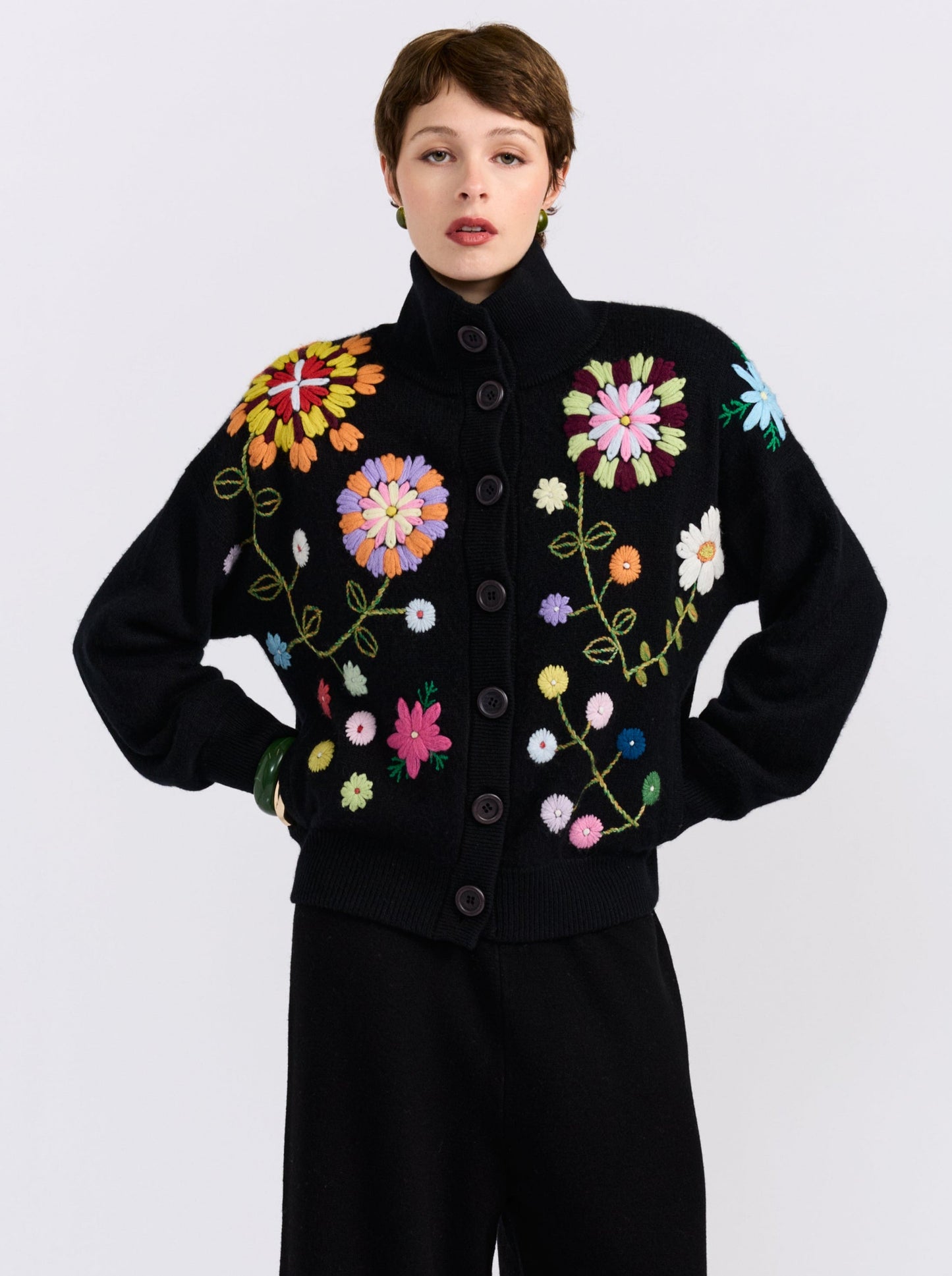 A model wearing a black knit bomber jacket with embroidered floral details, paired with black trousers, against a light gray studio background.

