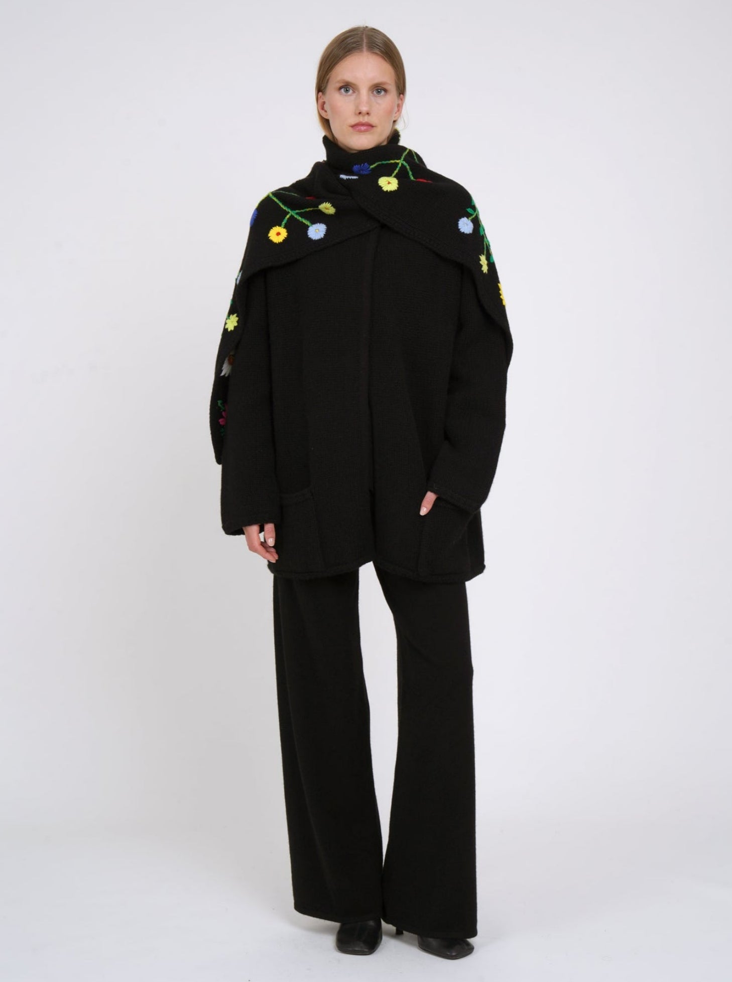 Black cashmere hip length jacket with long sleeves, patch pockets & flower embroidered detachable scarf wrapped closely around; worn with black cashmere trousers.
