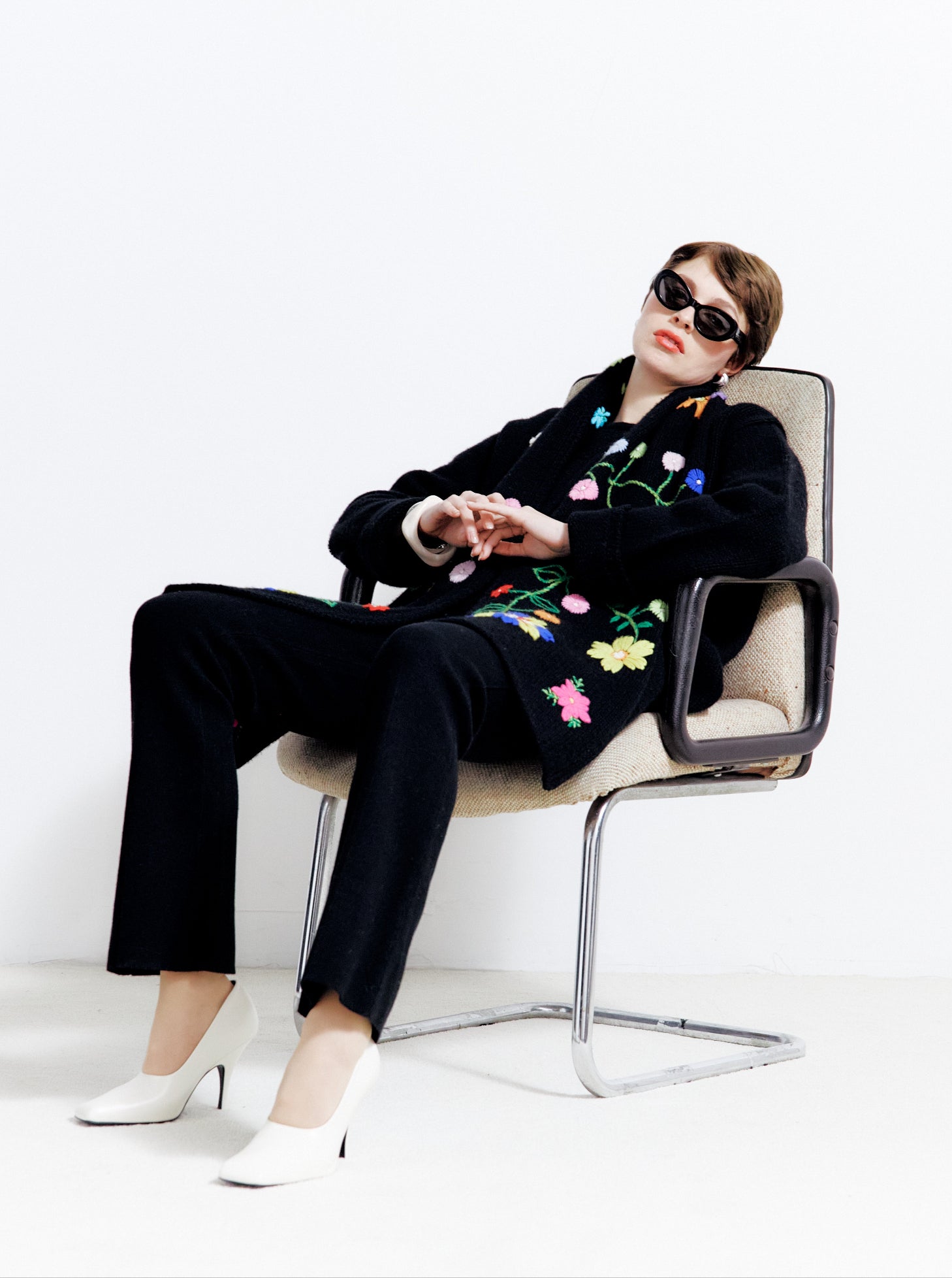 Seated model in black cashmere jacket with long sleeves, bright flower embroidered detachable scarf; worn with black cashmere trousers, black shades, white high heels.