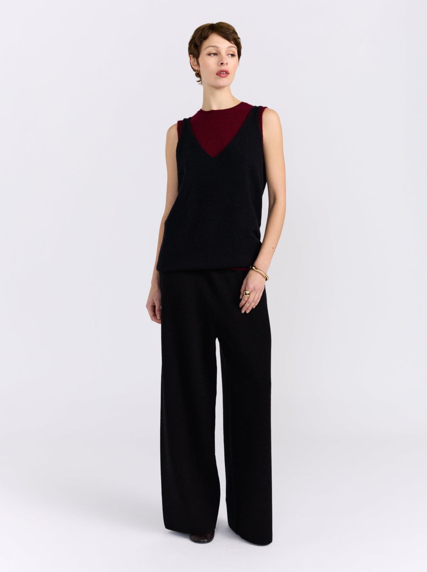 A model stands against a light grey studio background wearing a sleeveless black and burgundy cashmere double layer top paired with wide-leg black trousers.