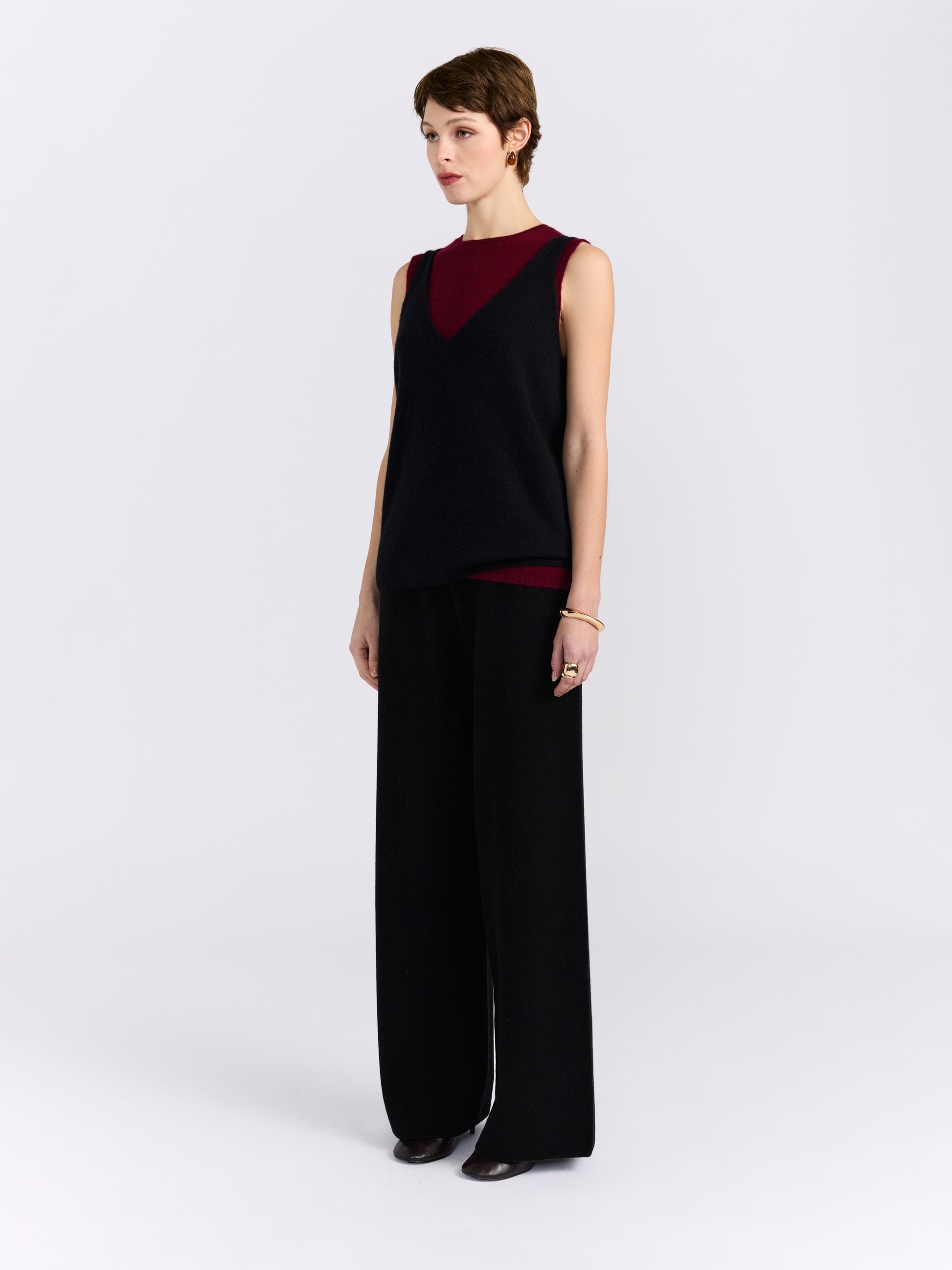 A model stands against a light grey studio background wearing a sleeveless black and burgundy cashmere double layer top paired with wide-leg black trousers.