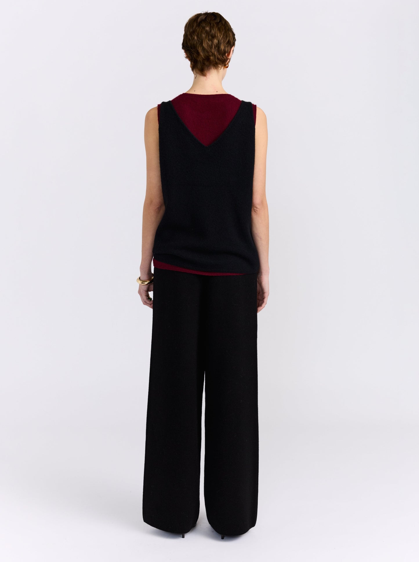 A model stands against a light grey studio background wearing a sleeveless black and burgundy cashmere double layer top paired with wide-leg black trousers.