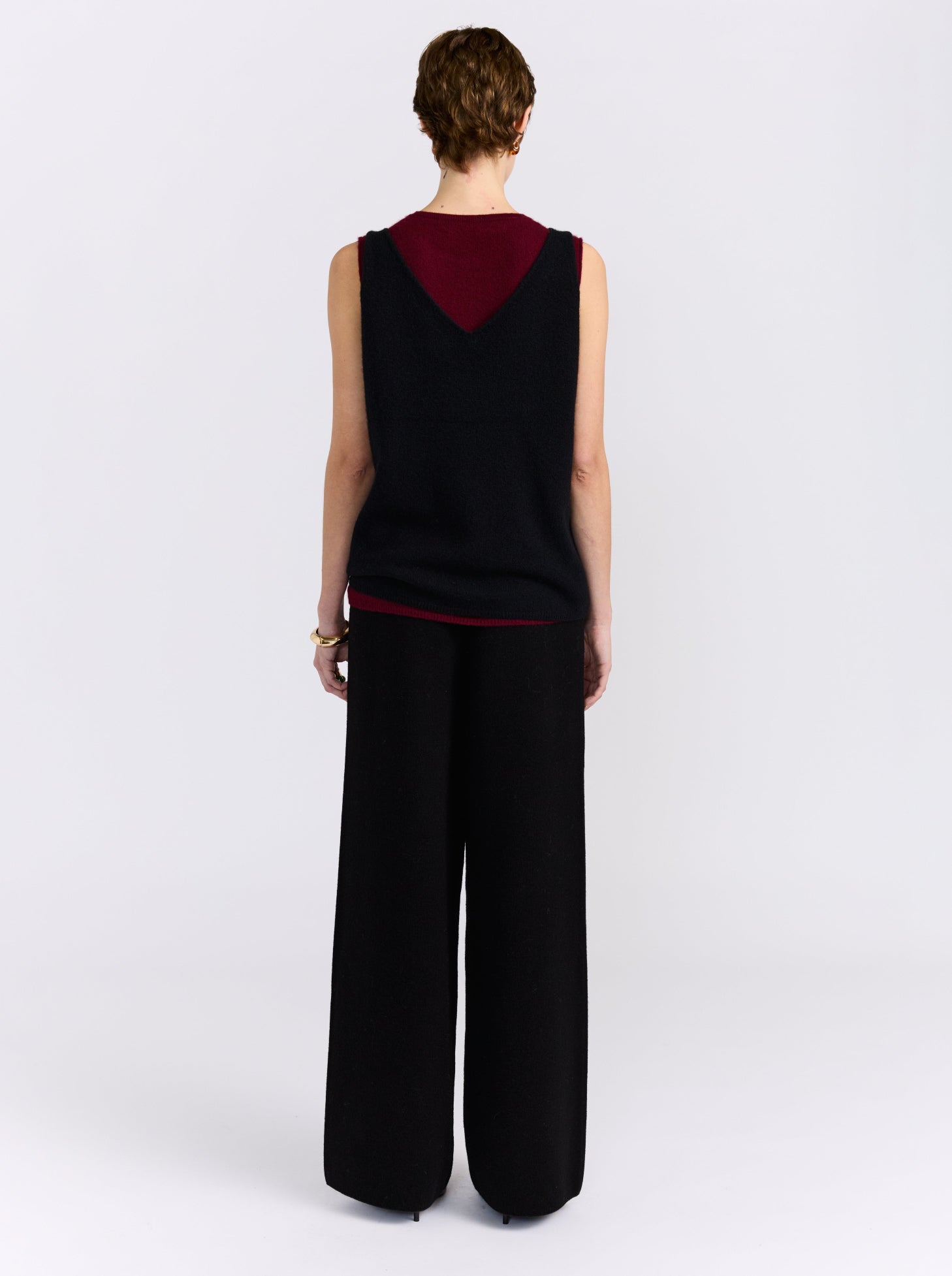 A model stands against a light grey studio background wearing a sleeveless black and burgundy cashmere double layer top paired with wide-leg black trousers.