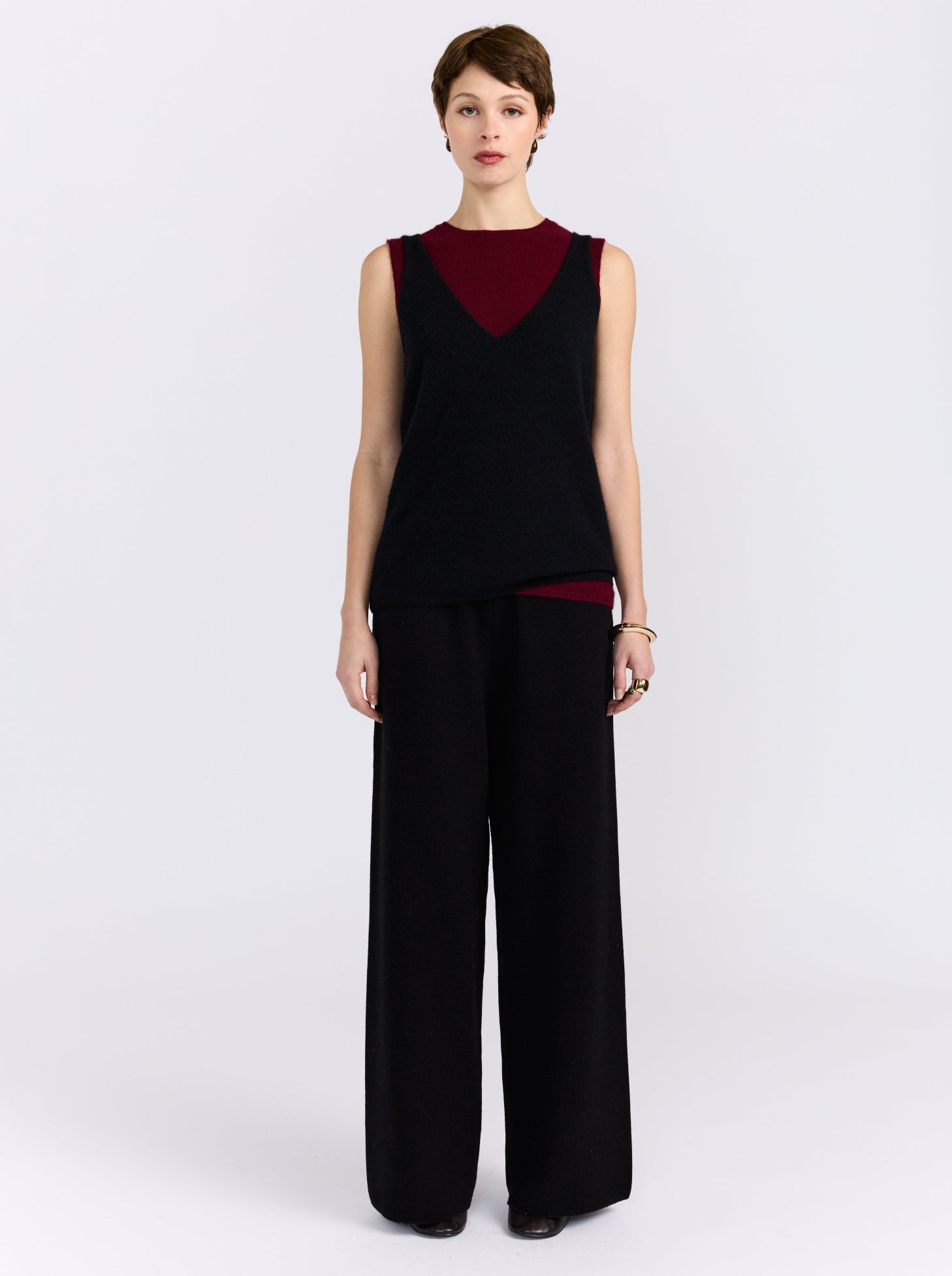 A model stands against a light gray studio background wearing a sleeveless black top with a deep V-neckline and a contrasting dark red inner neckline, paired with wide-leg black trousers.