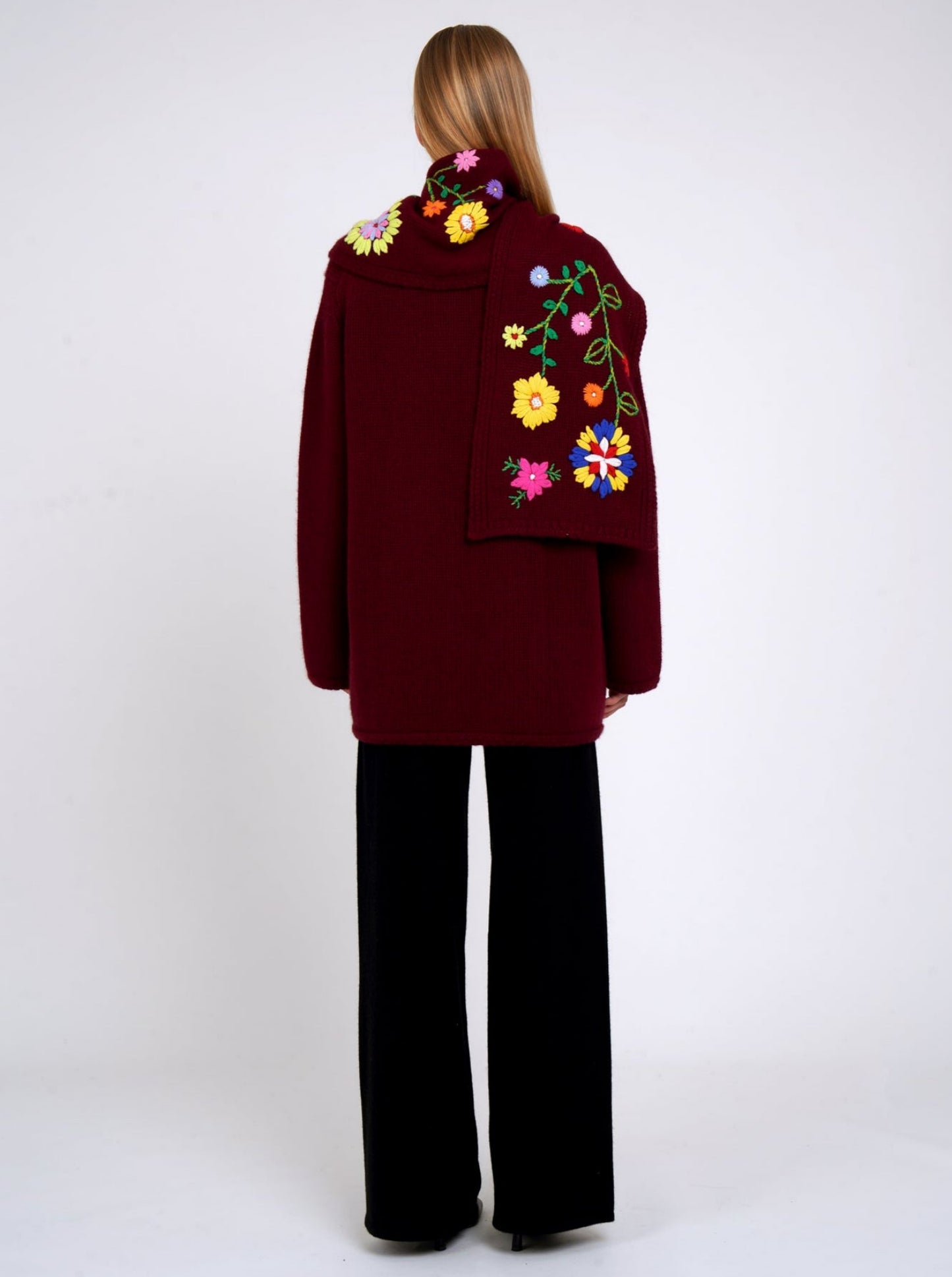 Back view burgundy cashmere hip length jacket with a colourful flower embroidered detachable scarf; worn with black cashmere trousers.