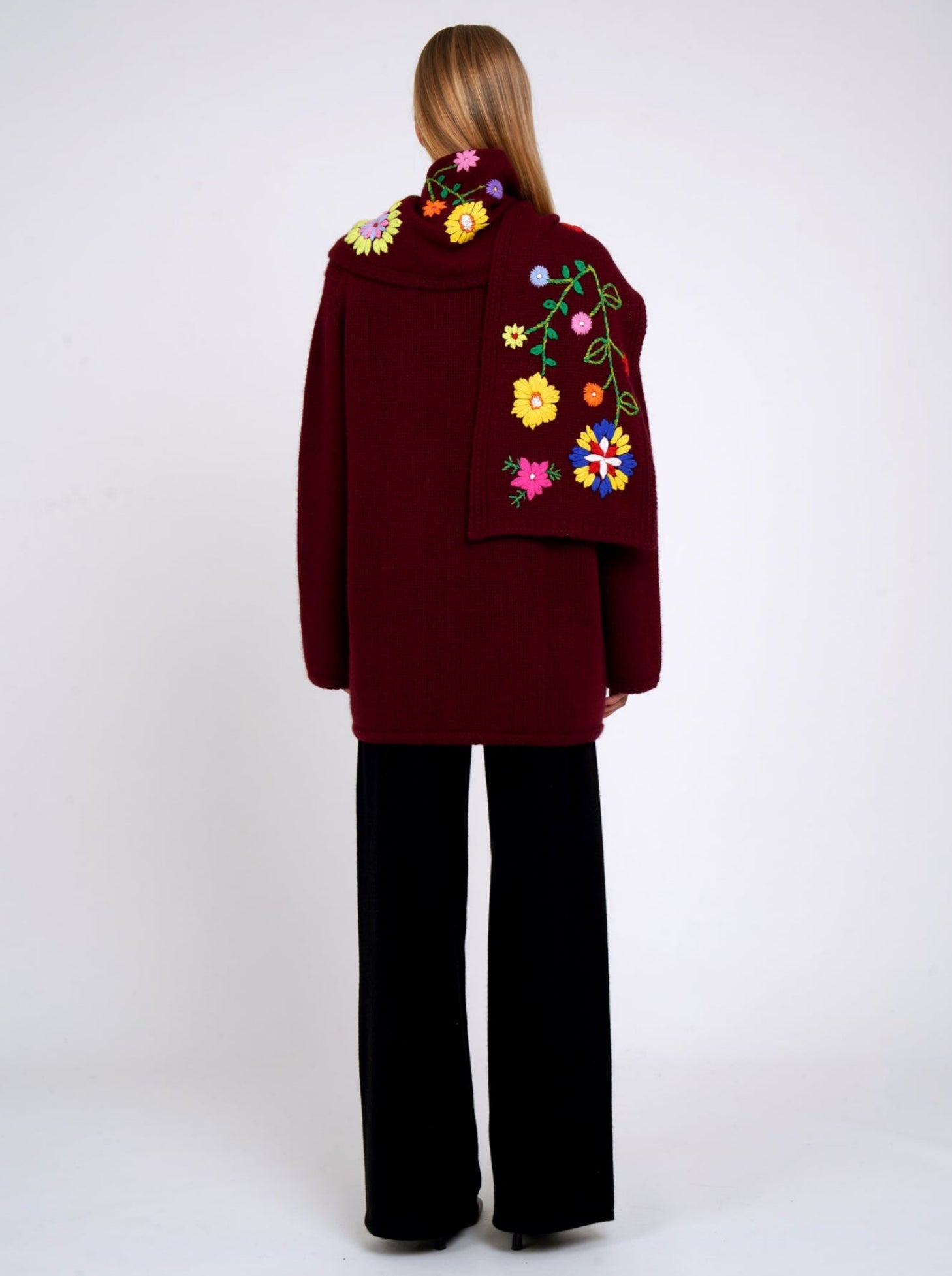 Back view burgundy cashmere hip length jacket with a colourful flower embroidered detachable scarf; worn with black cashmere trousers.