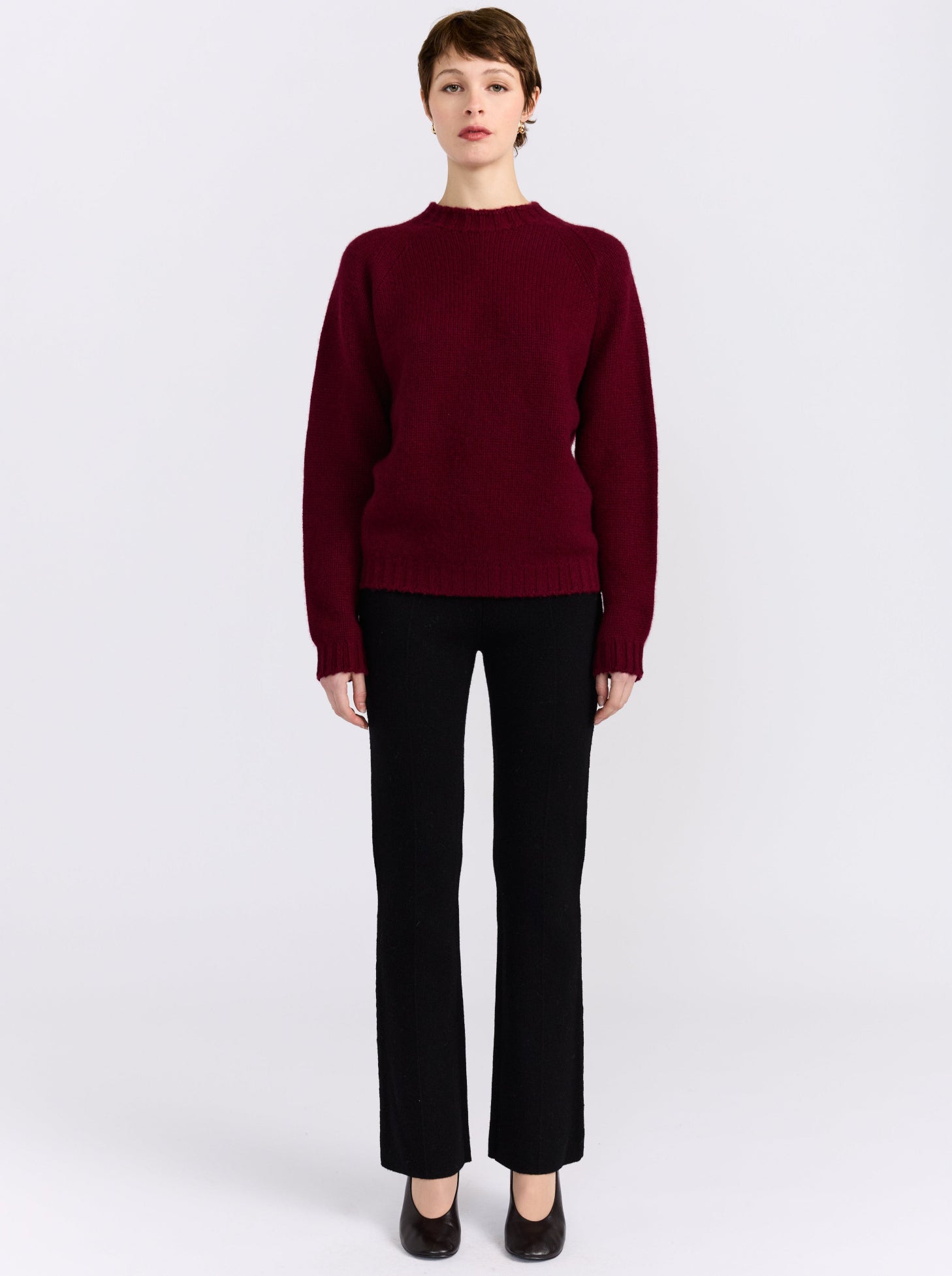 A model stands against a light gray studio background wearing a dark burgundy knit sweater, black trousers, and black shoes.