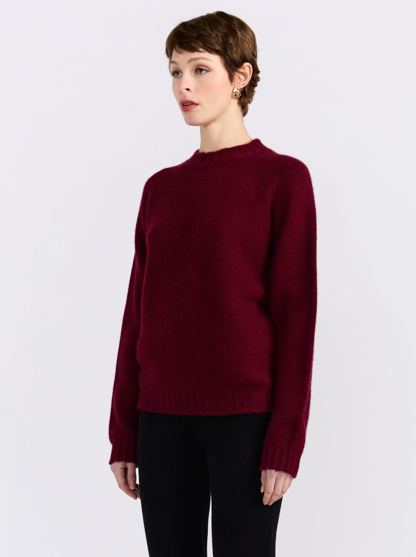 A model wearing a dark burgundy knit crewneck sweater and black pants, standing against a light gray studio background.