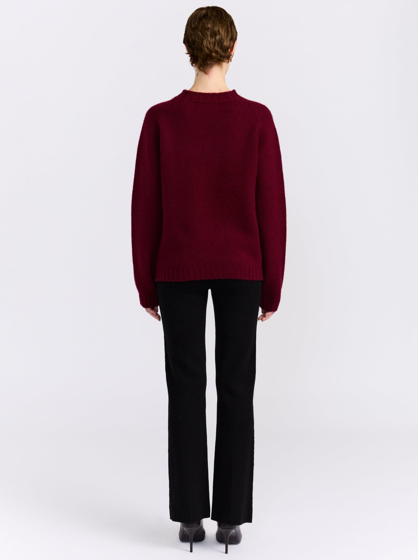 A model viewed from the back wearing a dark burgundy knit sweater and black trousers, standing against a plain light gray studio background.